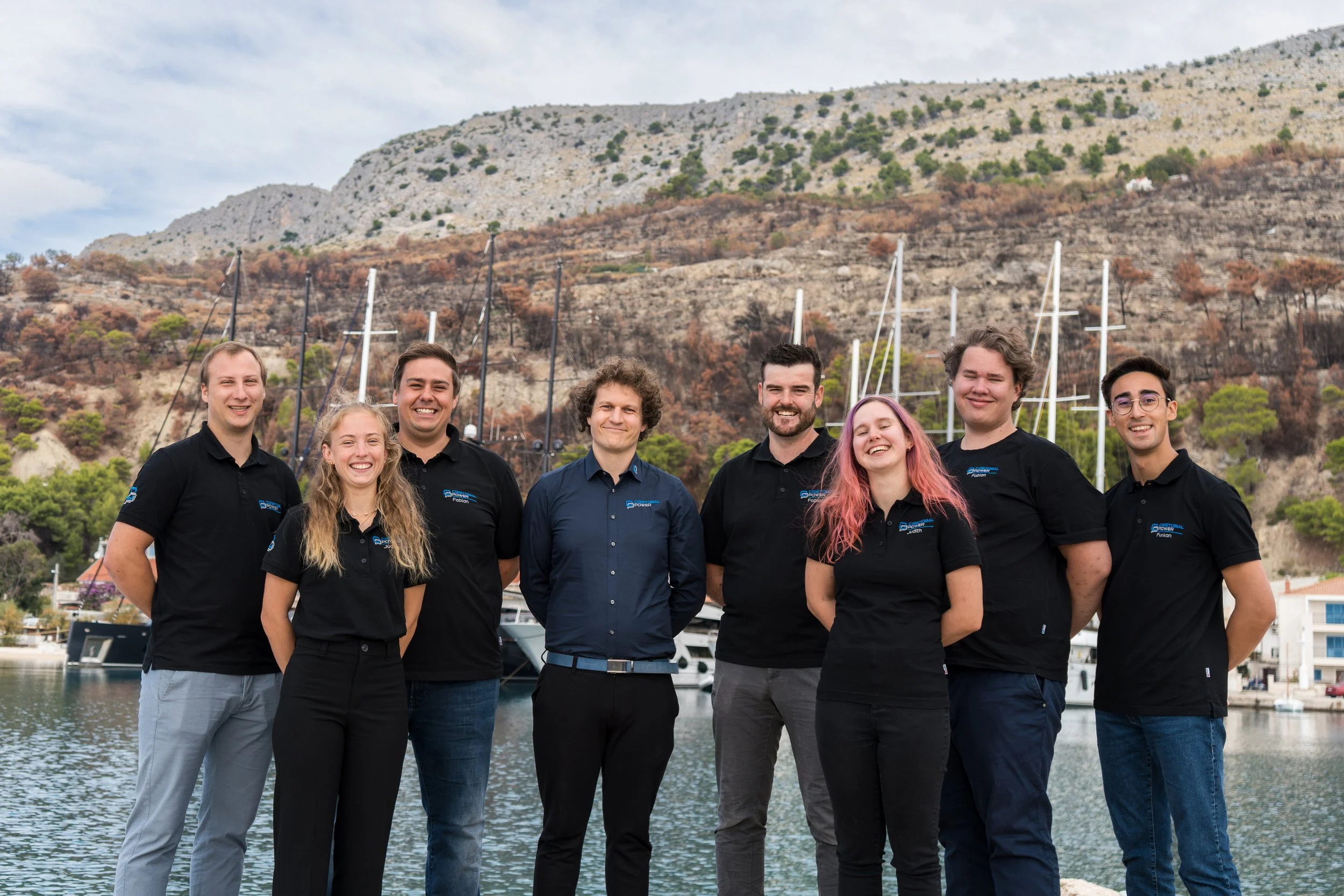 Group of eight young adults standing outdoors by a marina with sailboats, hills, and trees in the background, all wearing black or dark blue polo shirts.
