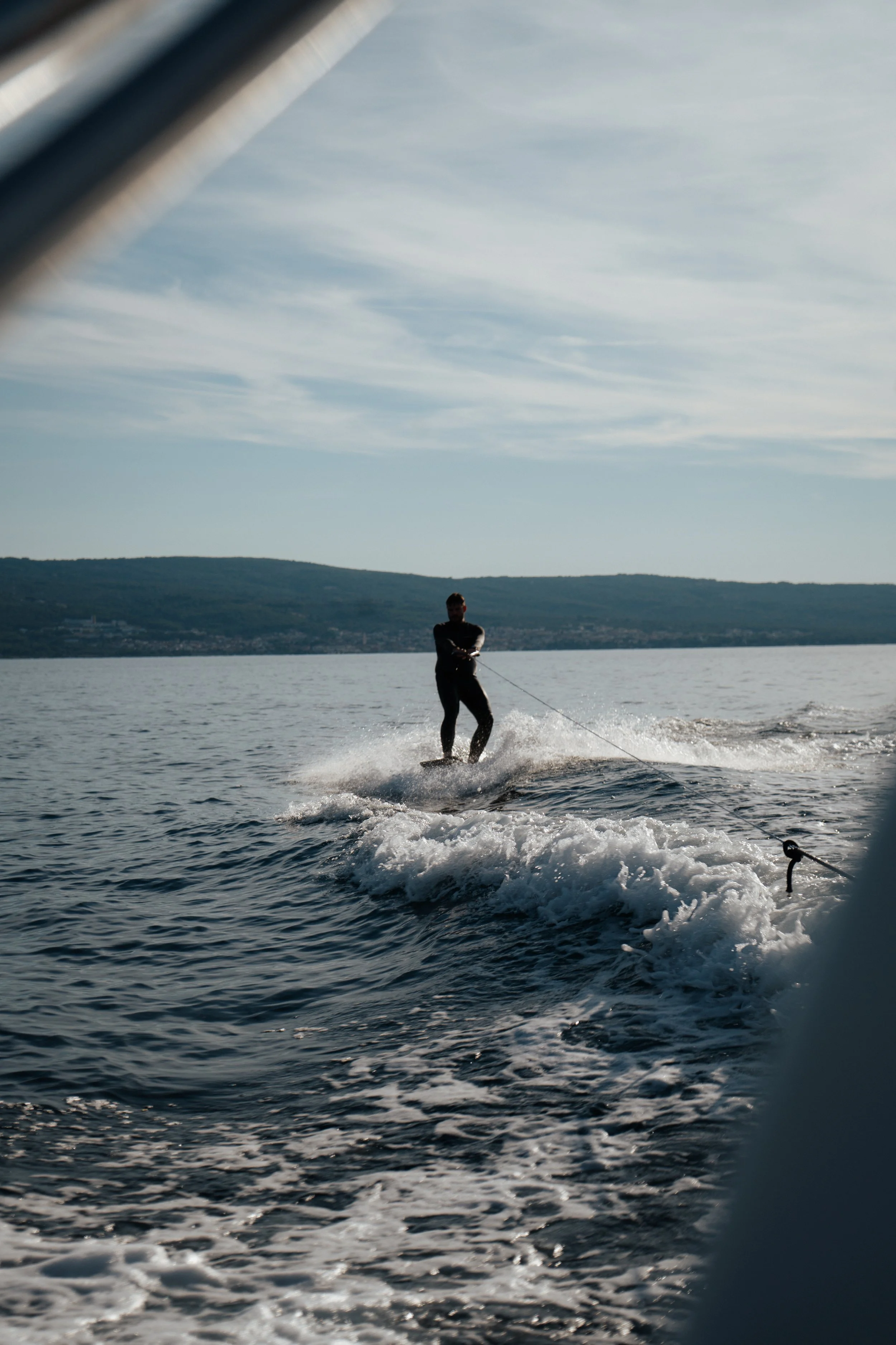 A person wakeboarding on a large body of water, with land and hills in the background, and a partially visible boat in the foreground.