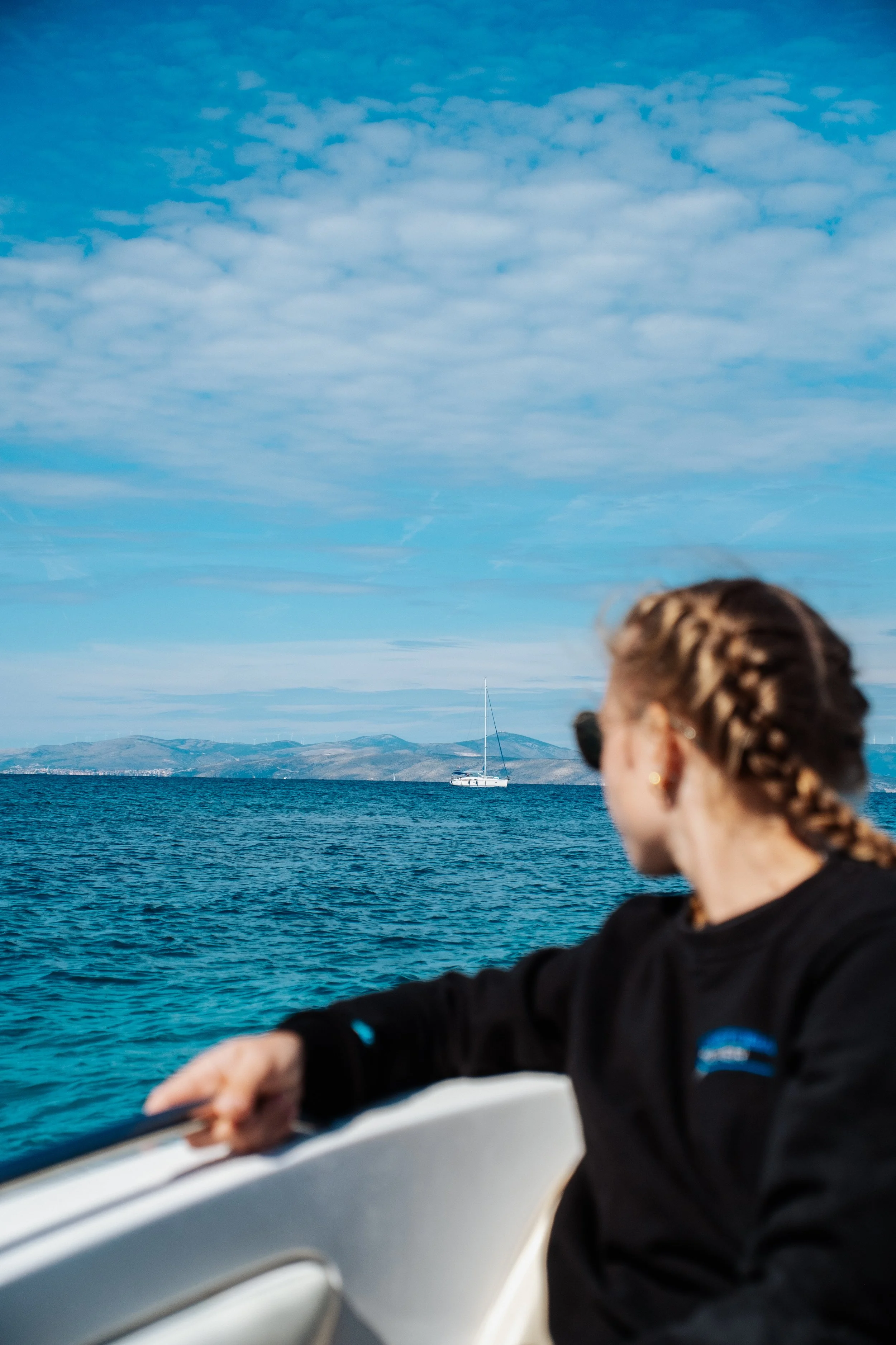 A woman with braided hair on a boat looking out at the ocean with a sailboat in the distance and hills on the horizon under a partly cloudy sky.