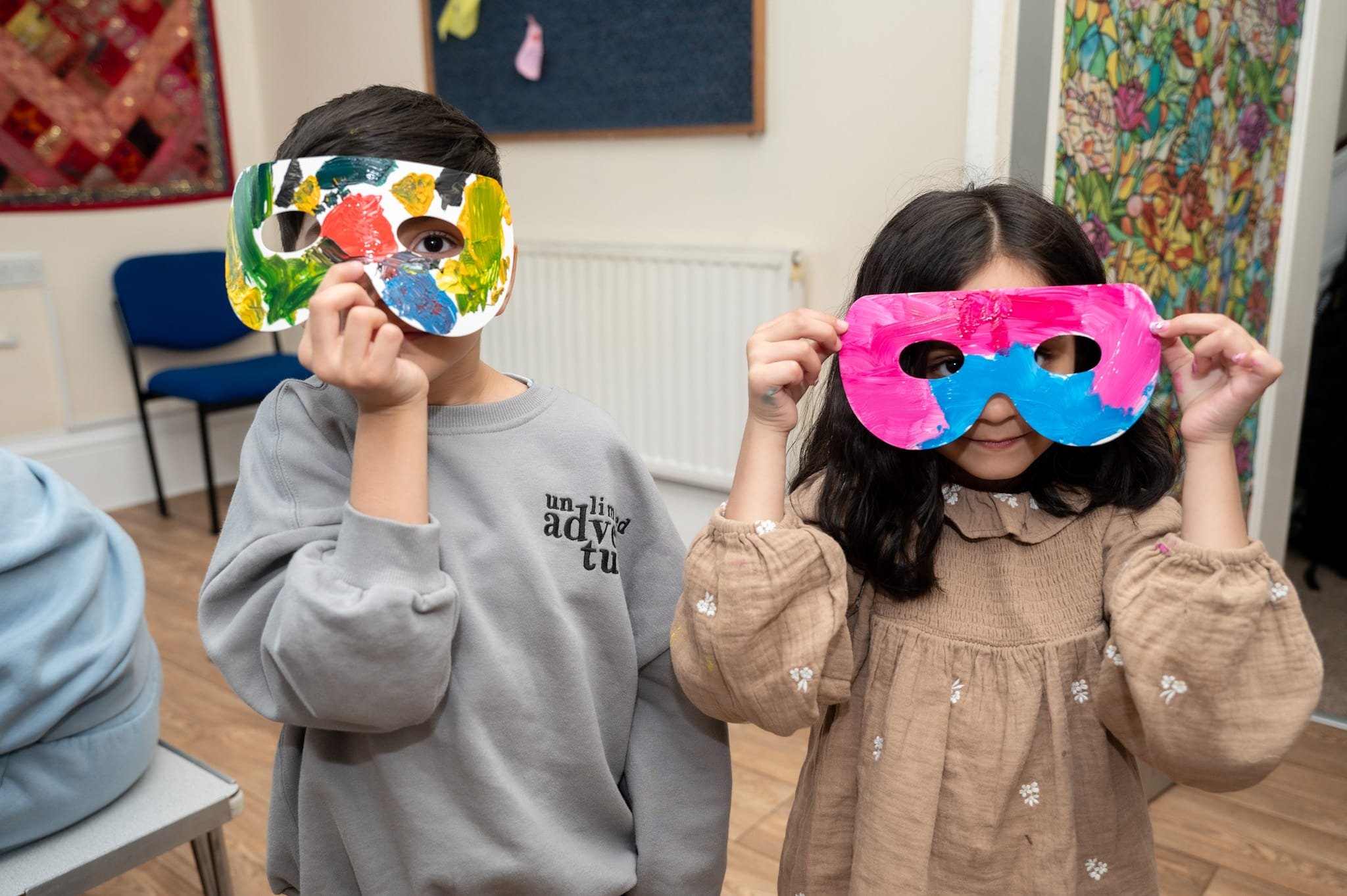 Two children holding colorful masks to their faces, standing in a room with wooden flooring and decorated walls.
