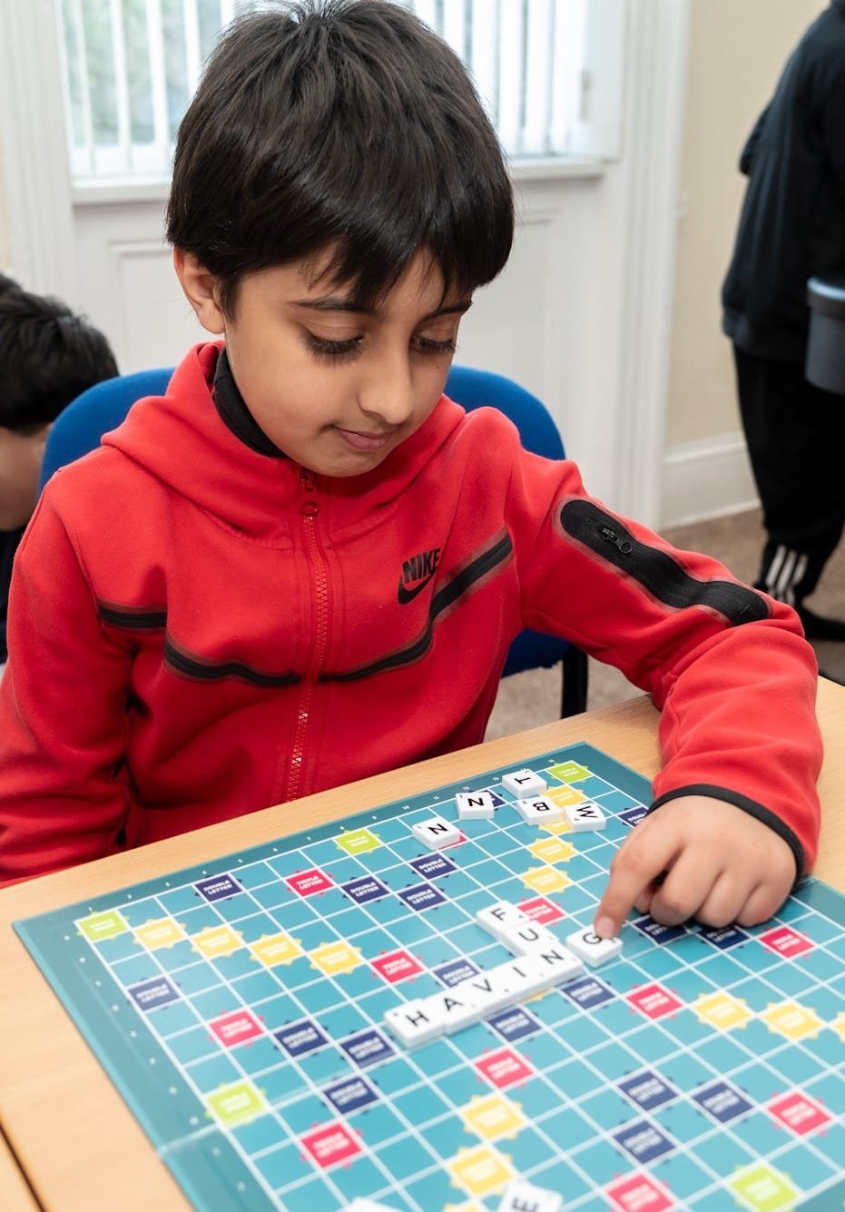 Child in red jacket playing Scrabble at a table, placing letter tiles on the board.