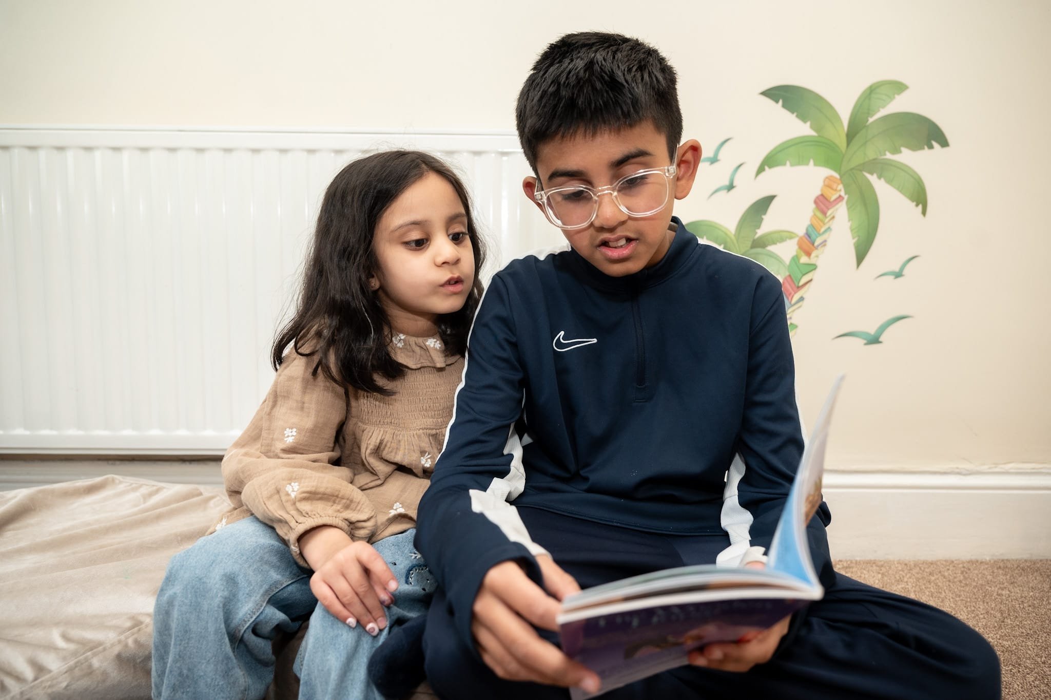 A young girl and an older boy sitting together on a cushion, with the boy reading a book aloud. The girl listens intently. They are in a room with a light-colored wall and a decorative palm tree decal.