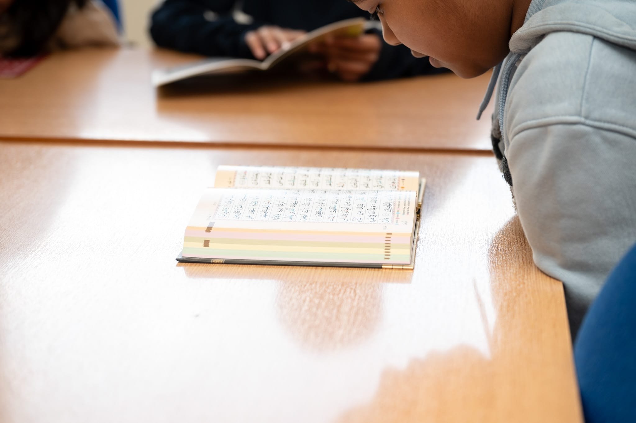 Child reading a book in Arabic script at a wooden table during a study session.