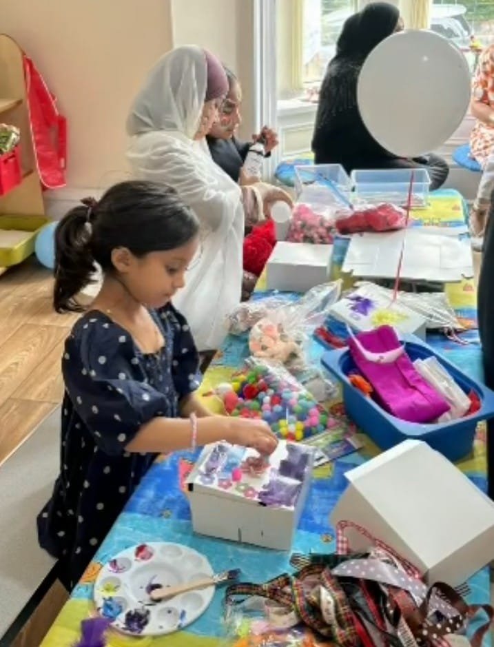 Children engaging in a craft activity at a table filled with art supplies, including paint, pom-poms, and ribbons, while adults assist them.