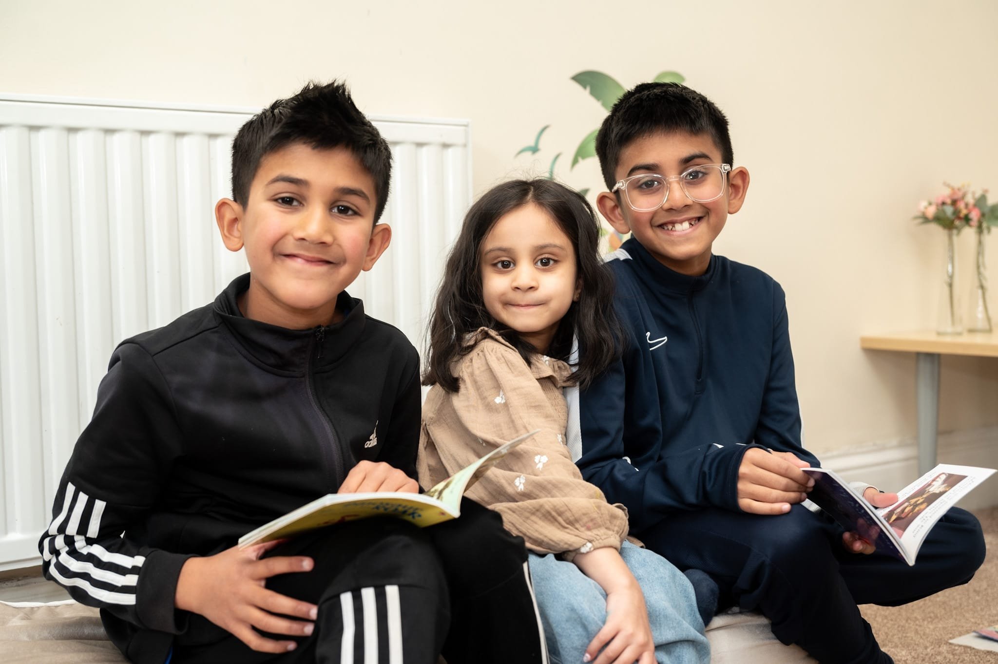 Three children sitting side by side, smiling and holding books. One child is wearing glasses. They are sitting indoors with a plant and vase in the background.