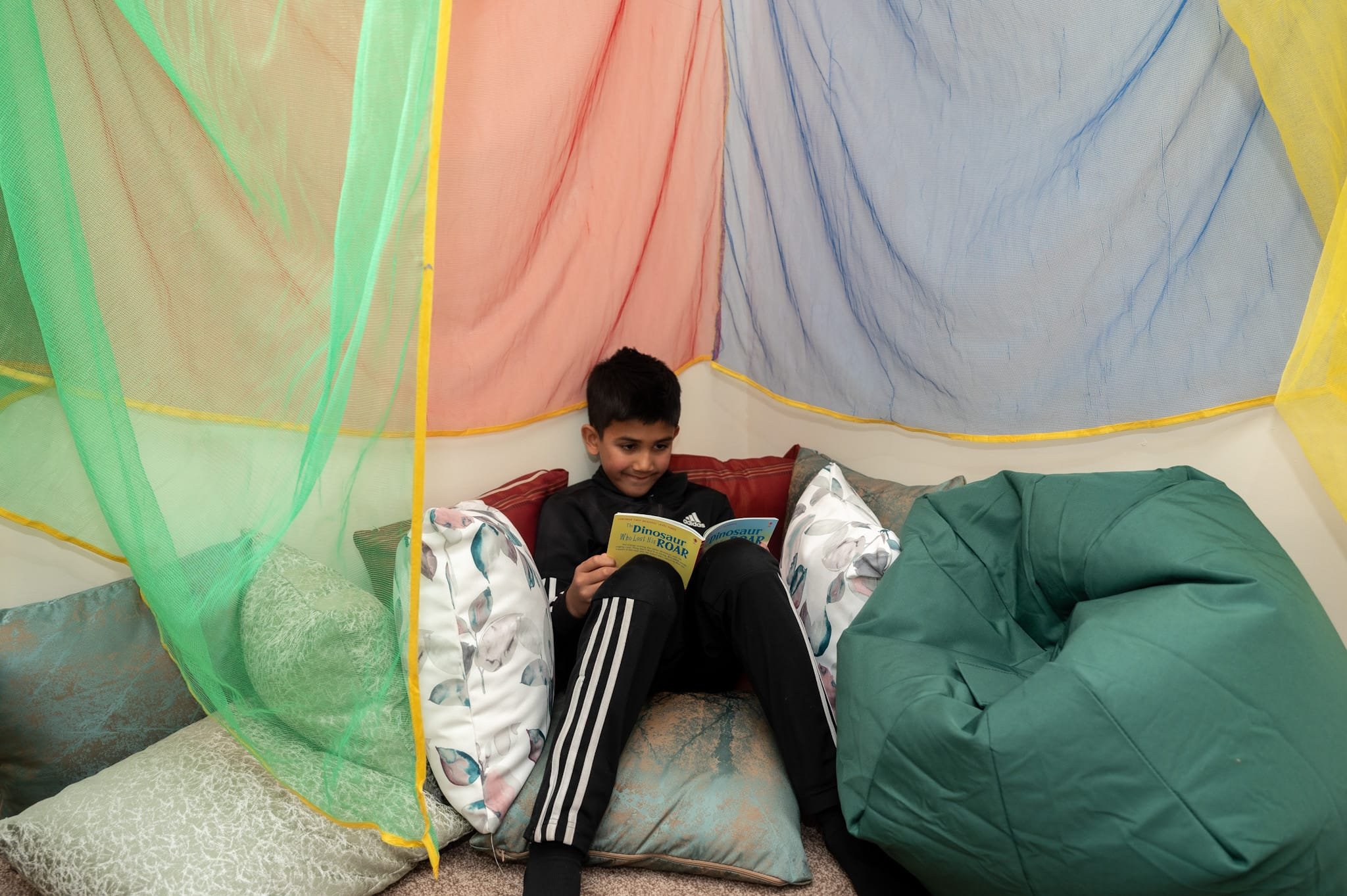 A child is sitting on a cushioned surface and reading a book titled 'Dinosaur Roar' inside a colorful blanket fort. The fort is made of green, red, blue, and yellow fabrics. A green beanbag is positioned nearby.