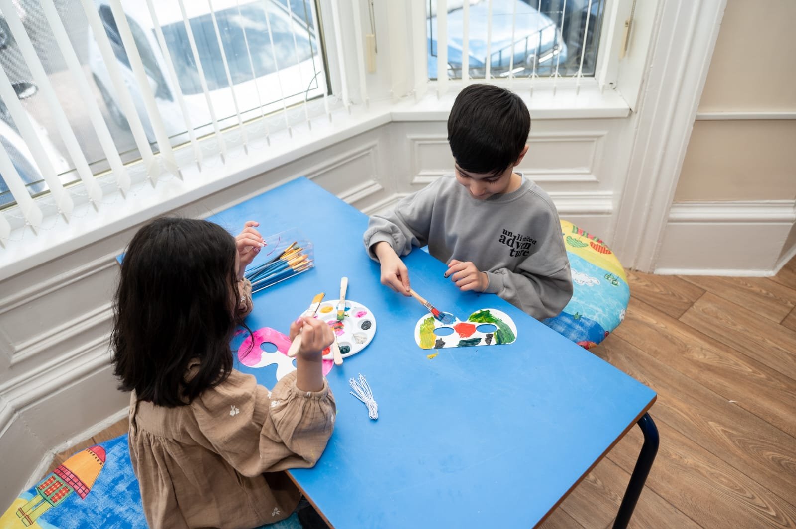 Two children painting at a table with brushes and palettes near a window.