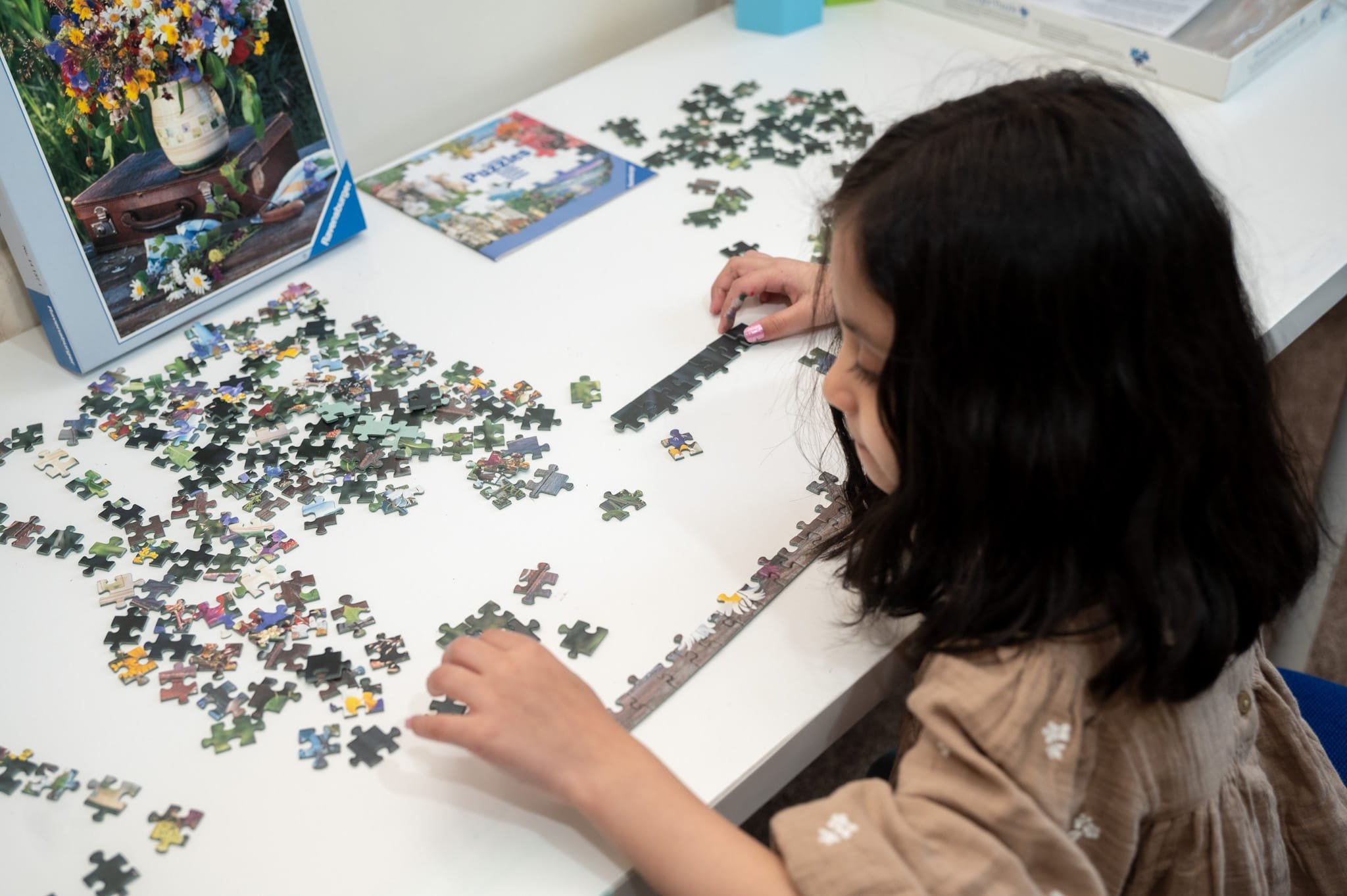 A child assembling a jigsaw puzzle on a white table, with puzzle box and pieces scattered around.