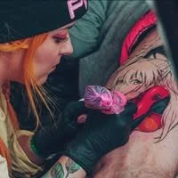Tattoo artist with orange hair and black gloves creating a colorful tattoo on a client's back.