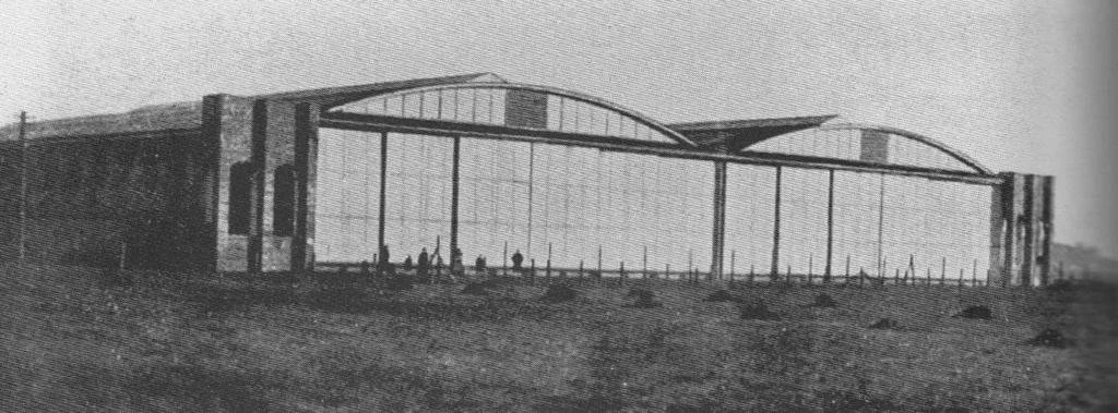 A vintage black and white photograph of a large greenhouse with a metal frame and glass panels, situated on a flat, open landscape.
