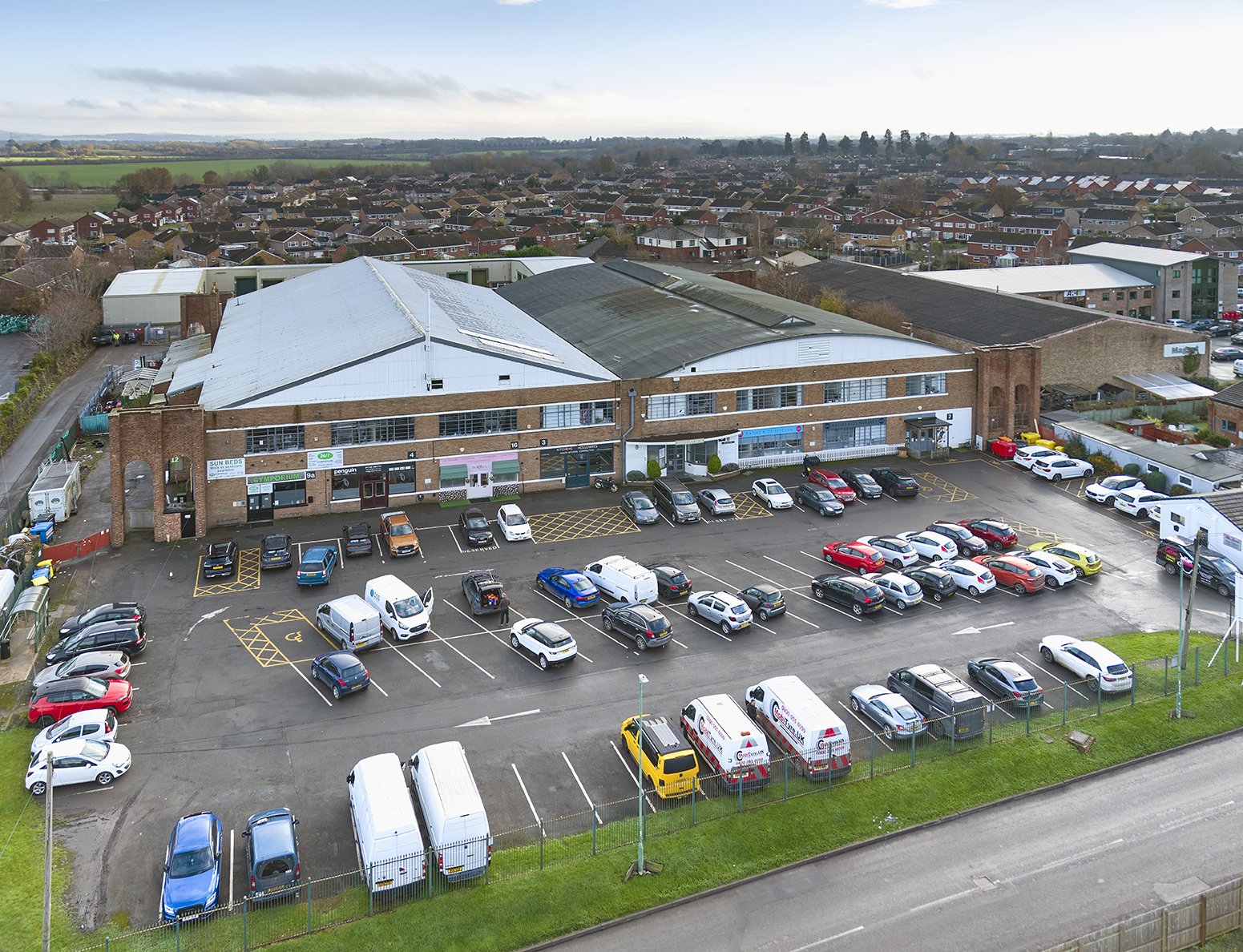 An aerial view of a parking lot in front of a commercial building with a suburban neighborhood and fields in the background.