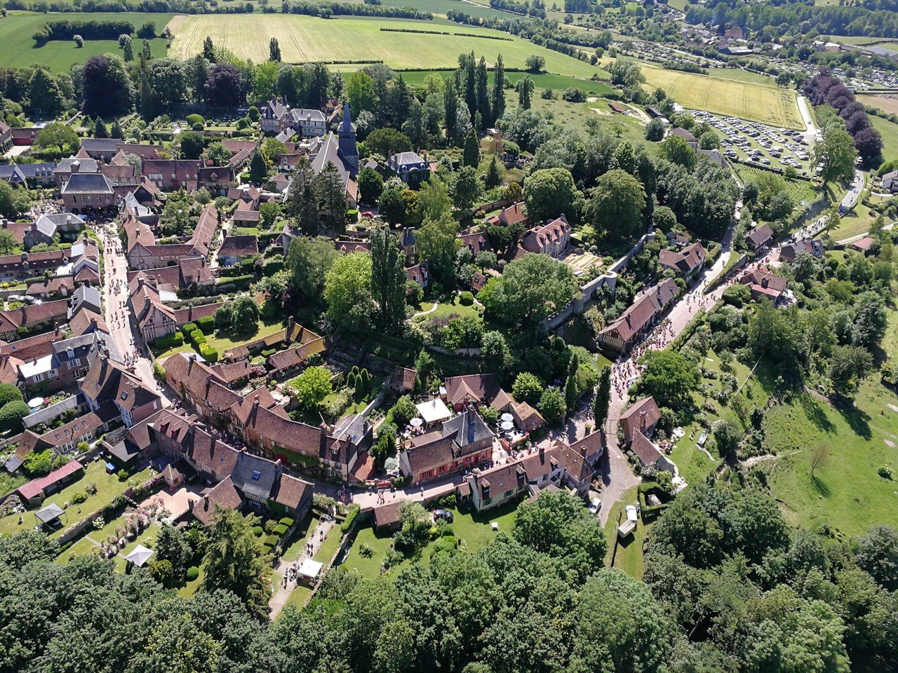 Vue aérienne d'un village pittoresque entouré de verdure, avec des maisons traditionnelles aux toits en tuiles rouges et des routes sinueuses. On aperçoit des champs et des arbres en arrière-plan.