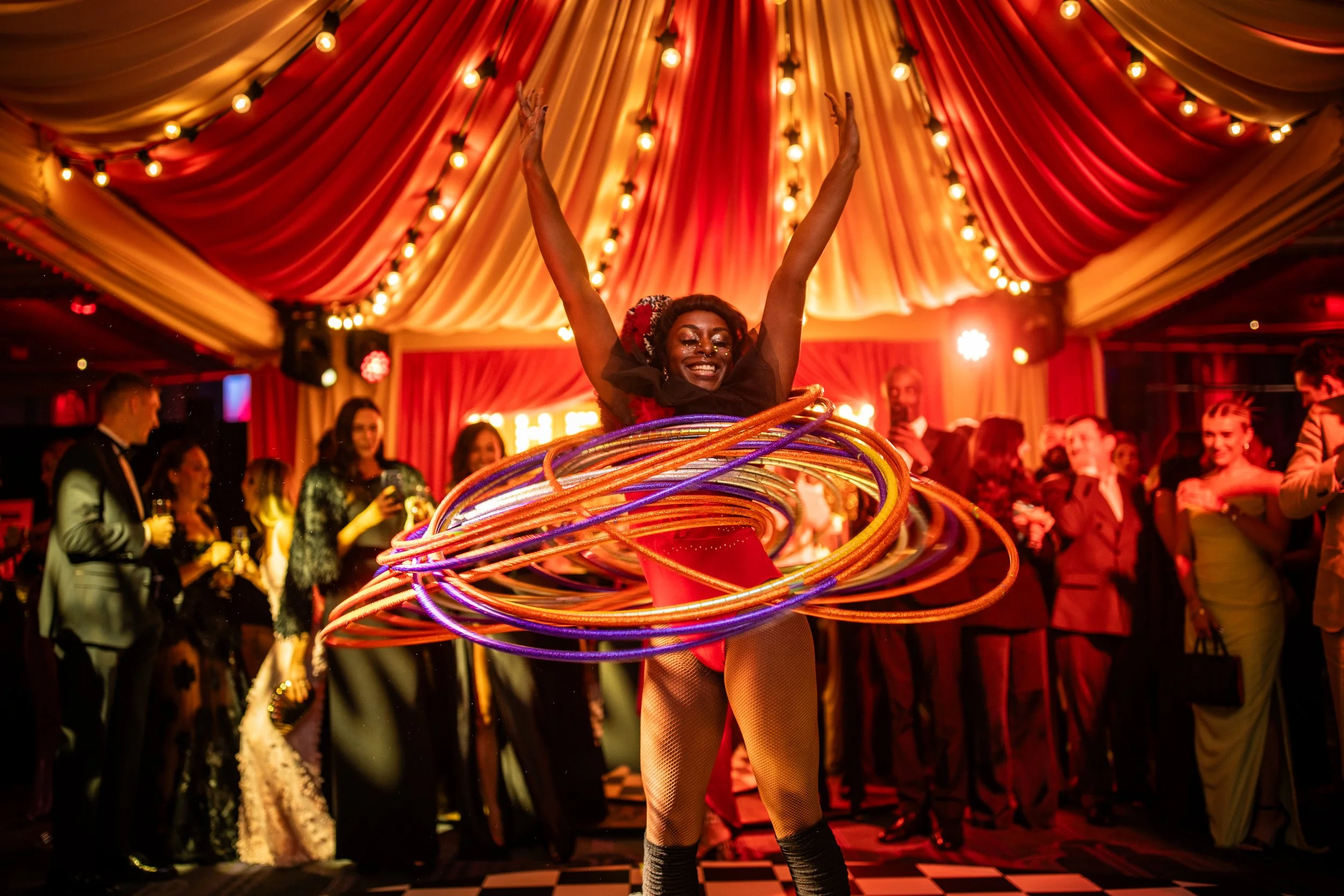 A woman performing a hula hoop routine at a lively indoor event, with warm, colorful lighting and an audience in the background.
