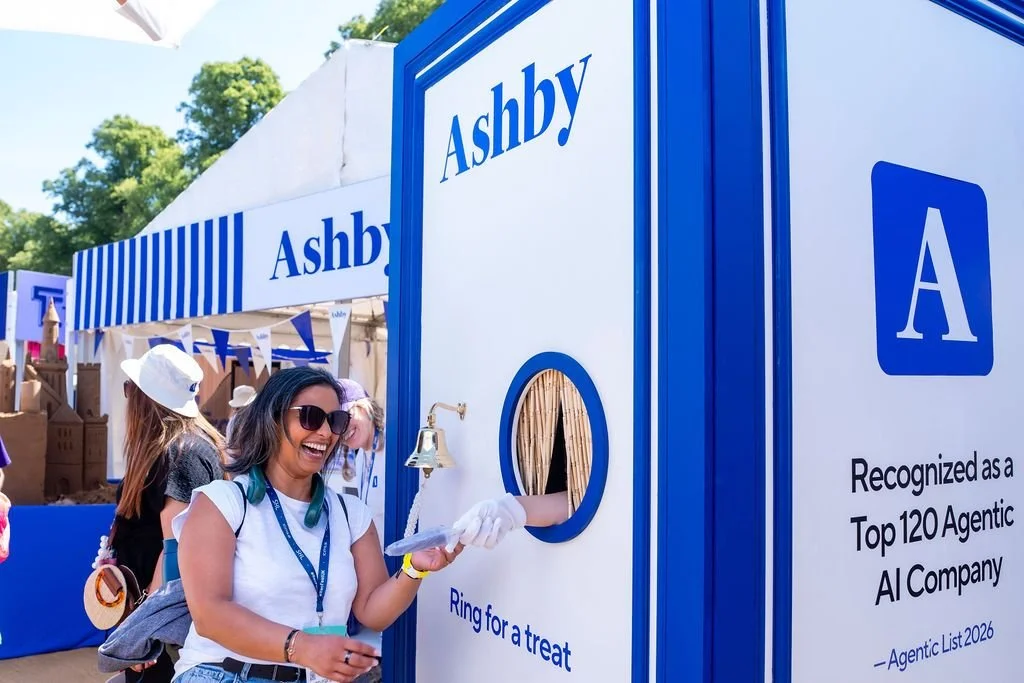 A woman at an outdoor event laughing and ringing a bell at a blue and white booth labeled 'Ashby,' which is recognized as a top AI company, with other people and trees in the background.