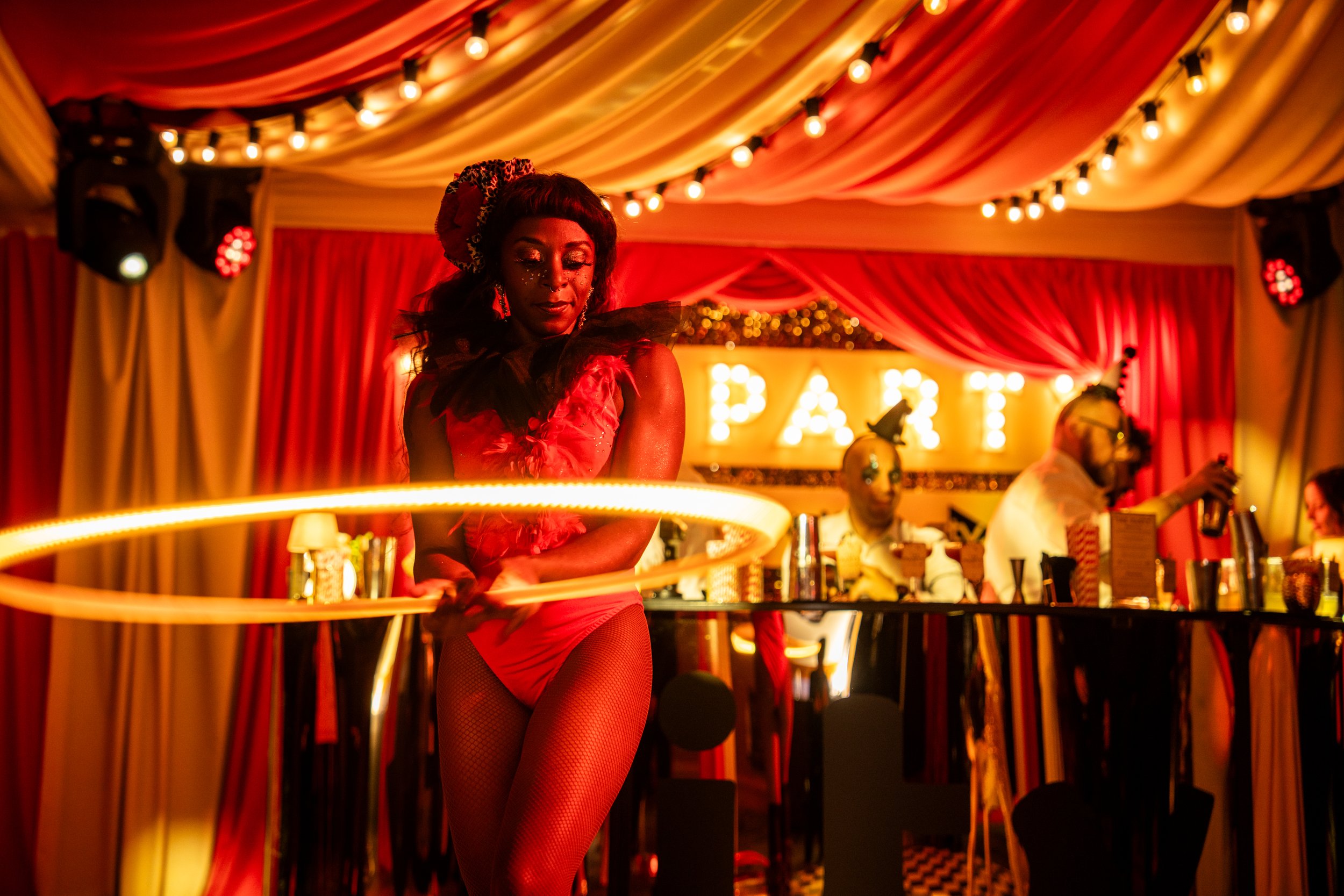 A woman dancing with a hoop in a circus or carnival tent decorated with red and yellow drapes and string lights. Other people are seated at a table in the background.