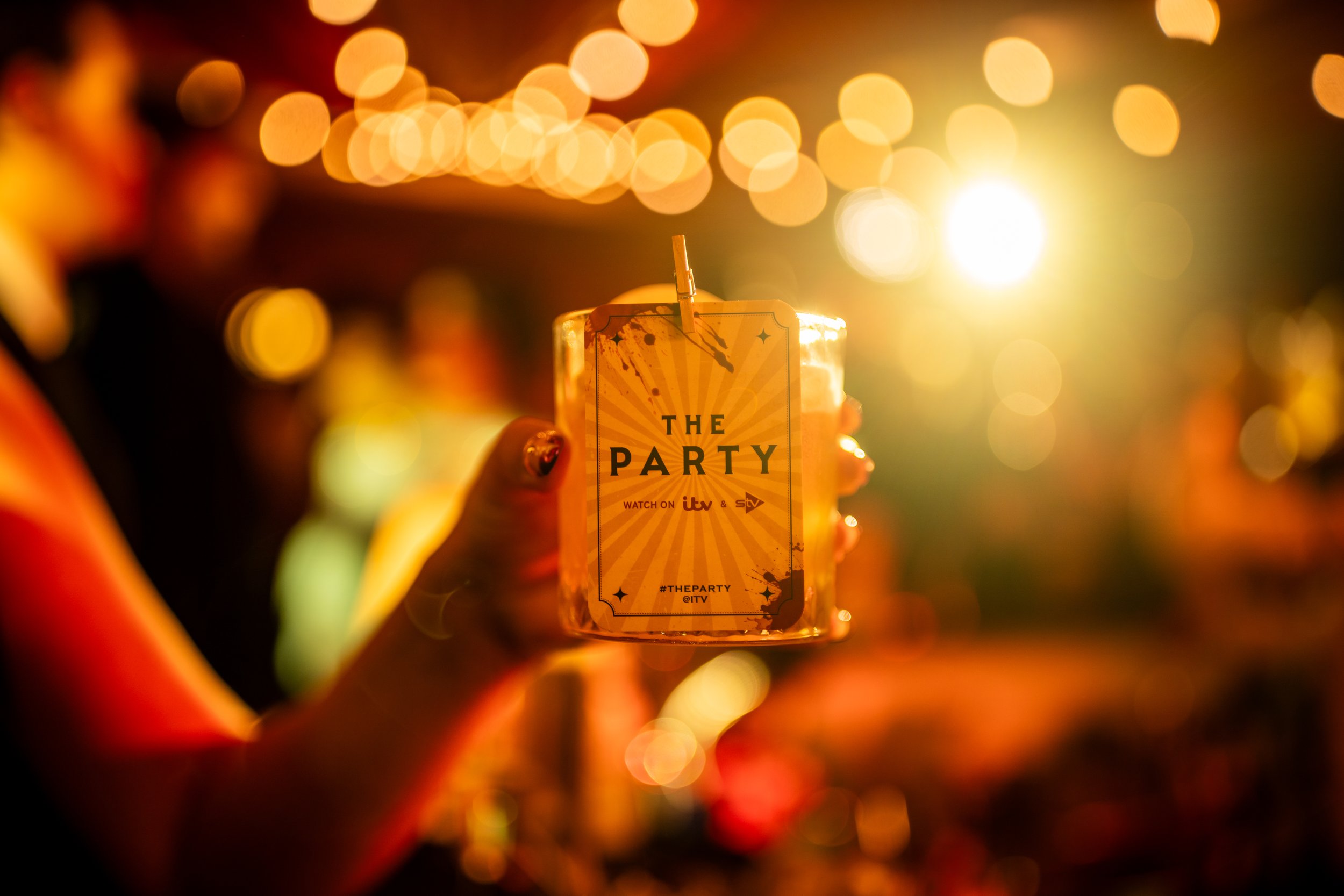 A hand holding a ticket with the text 'The Party' at a lively event with warm lighting and bokeh background.