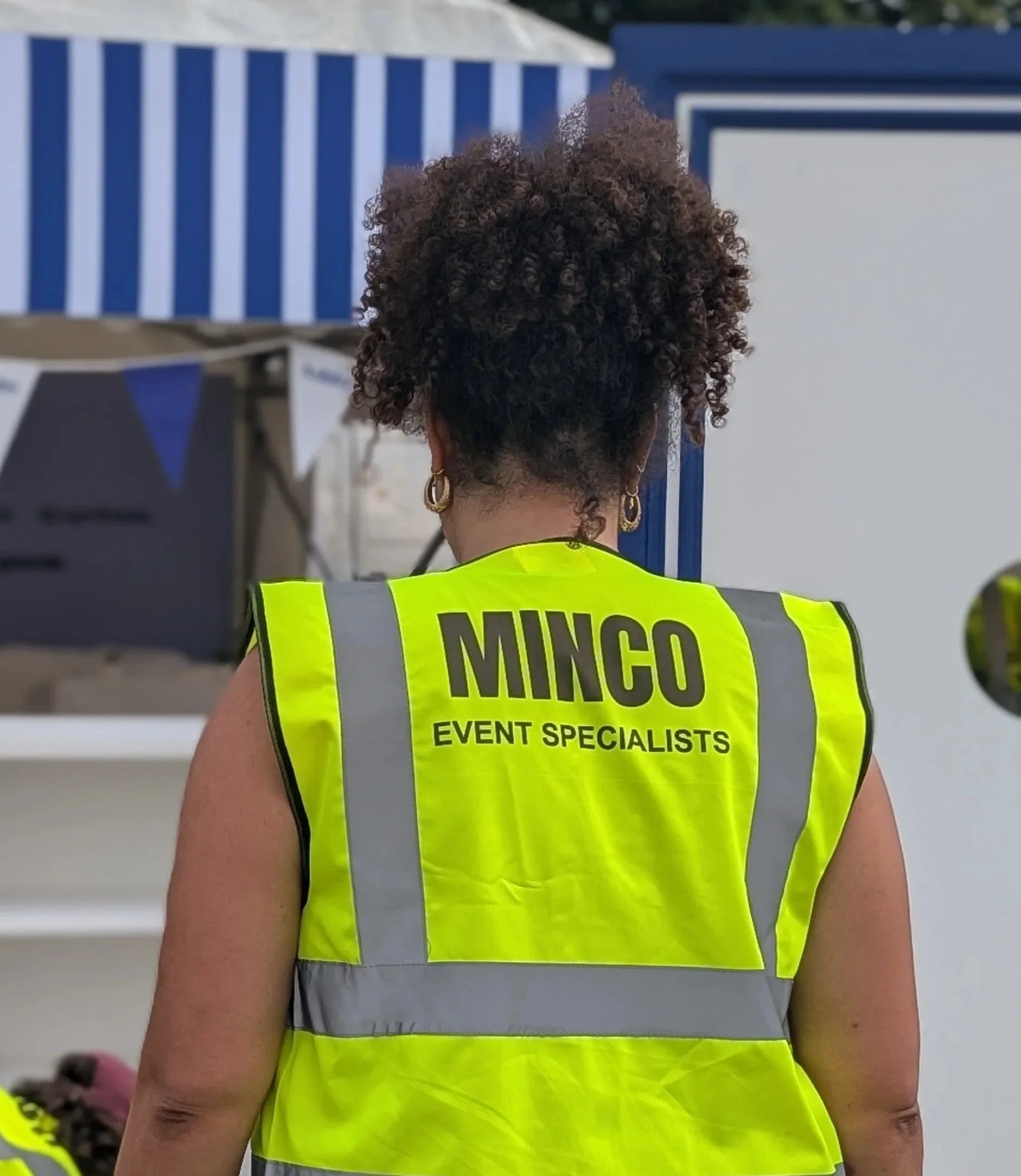 Person wearing a neon yellow safety vest with the text 'MINGO EVENT SPECIALISTS' on the back, and gold hoop earrings, with curly brown hair, at an outdoor event.