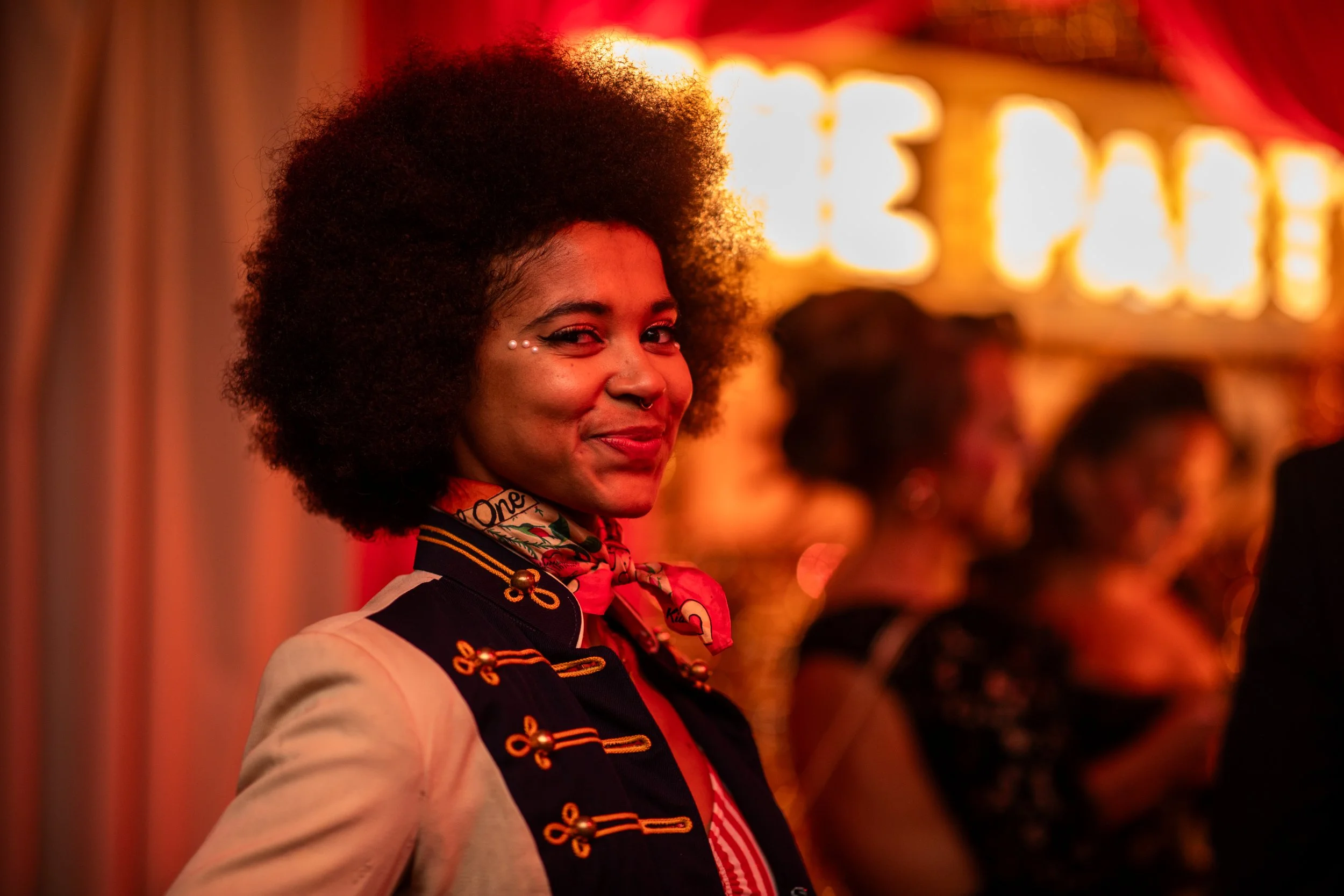 A woman with curly hair smiling, wearing a vintage-style jacket with gold embellishments, a colorful scarf, and makeup with decorative dots near her eye, standing in a dimly lit environment with bright orange lights and people in the background.