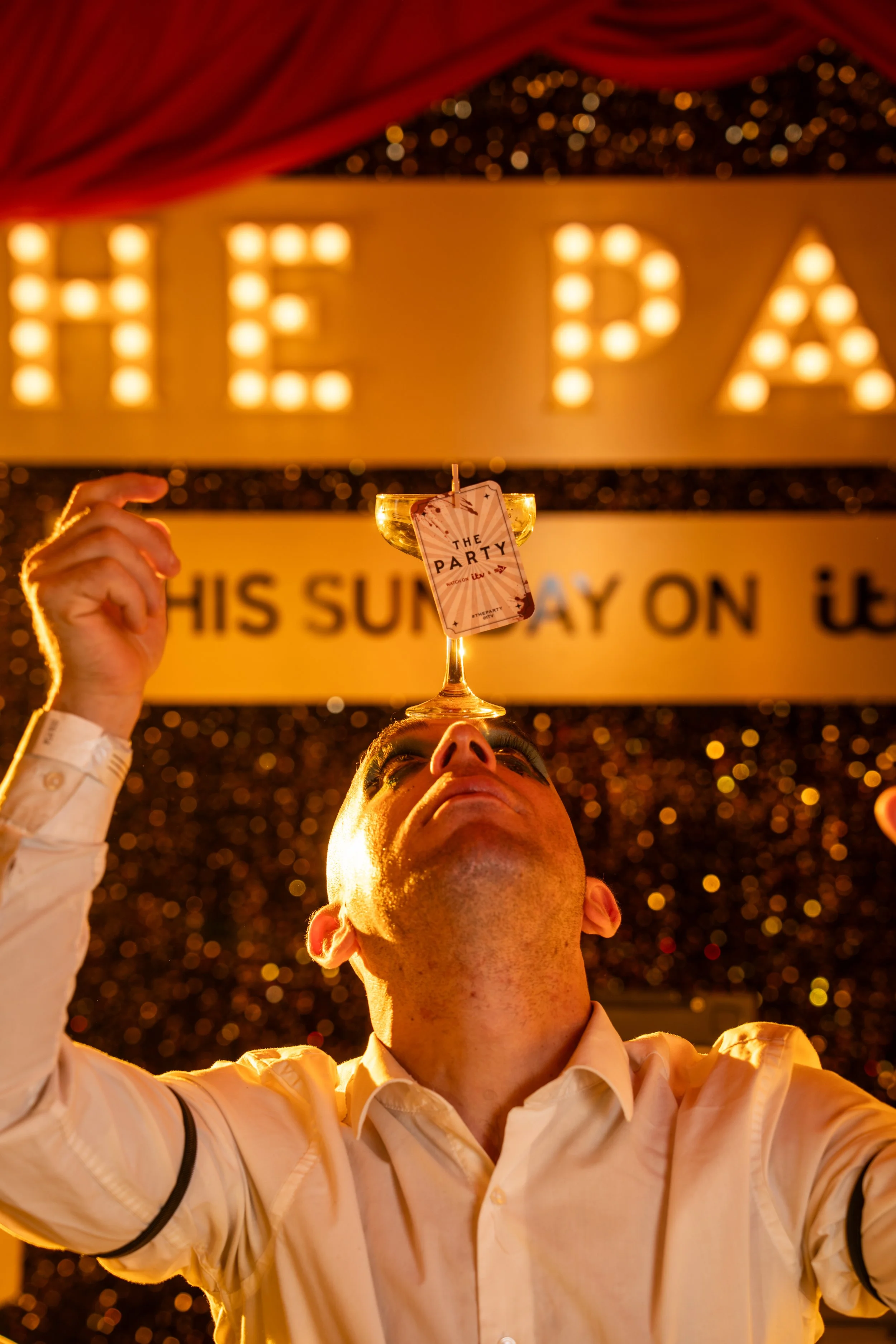 A person holding a lit champagne glass with a party tag that reads 'The Party' in front of a brightly lit sign spelling 'HELP' at a celebration event.