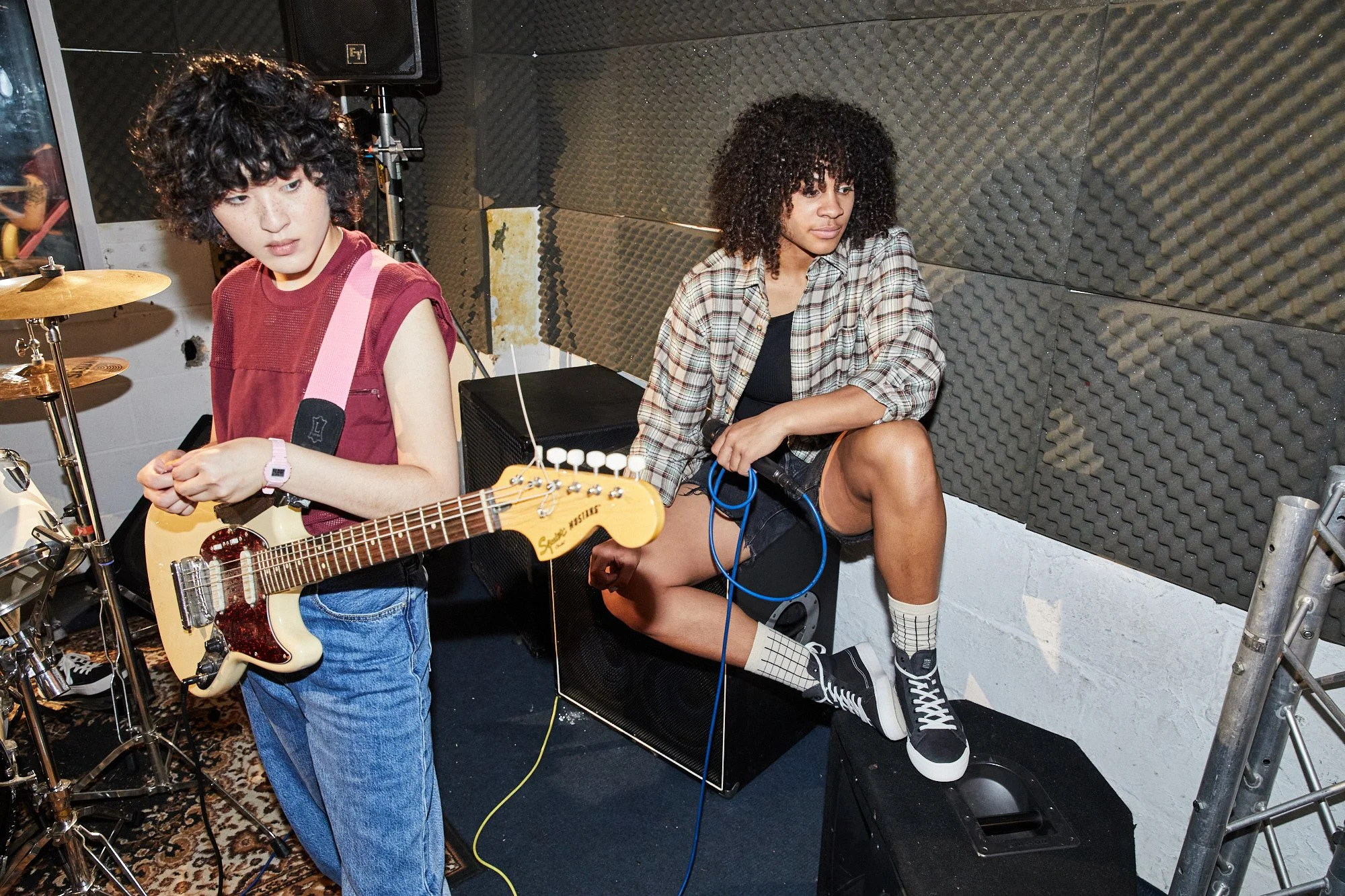Two women in a music rehearsal room with soundproof foam panels. One woman with curly hair is sitting on the floor holding a microphone, and the other woman with afro hair is sitting on a chair, wearing plaid shirt and holding a cable.