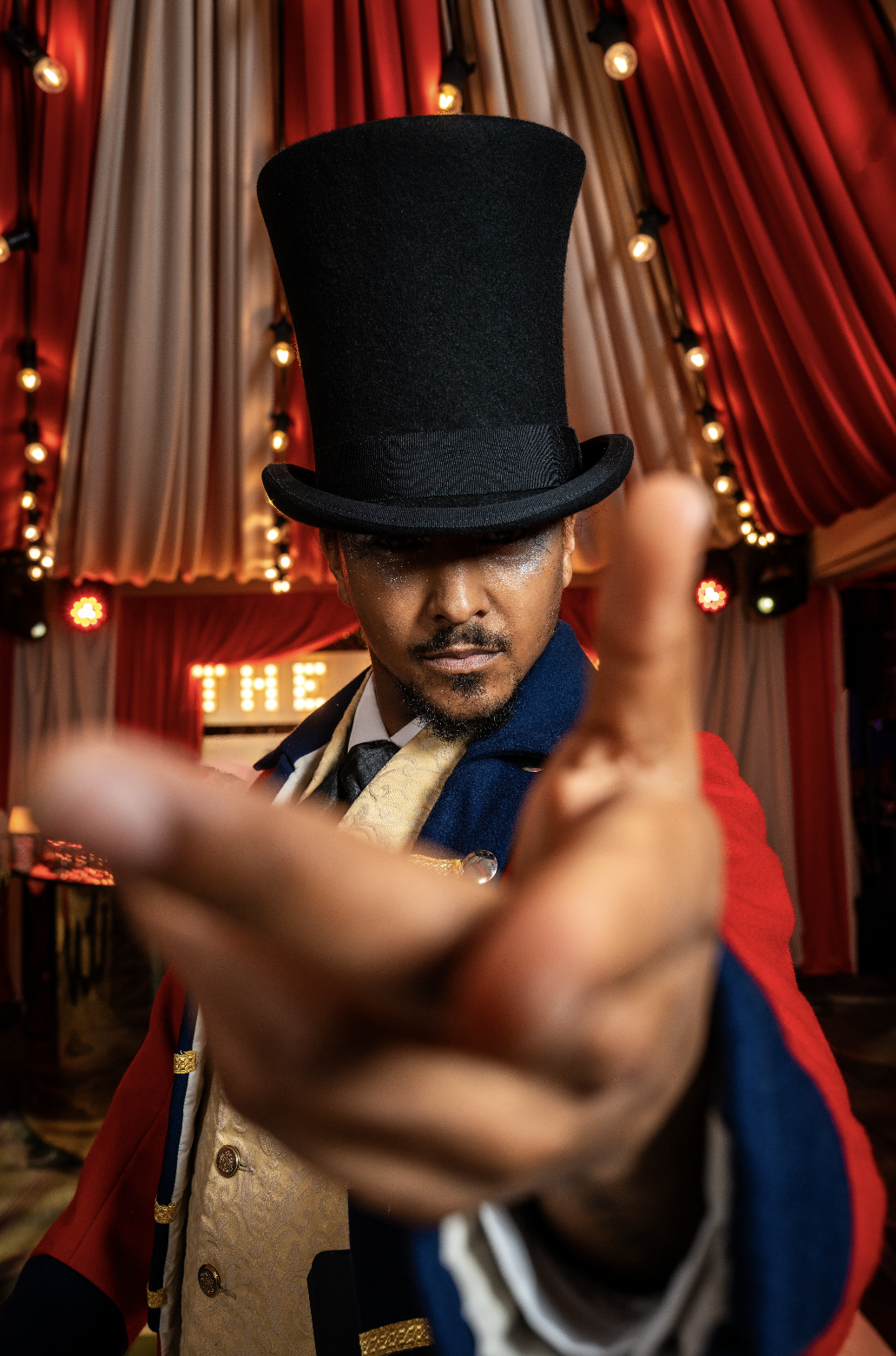 A man dressed as a circus or carnival performer wearing a tall black top hat and colorful costume, reaching towards the camera with a dramatic background of red and beige curtains and stage lights.