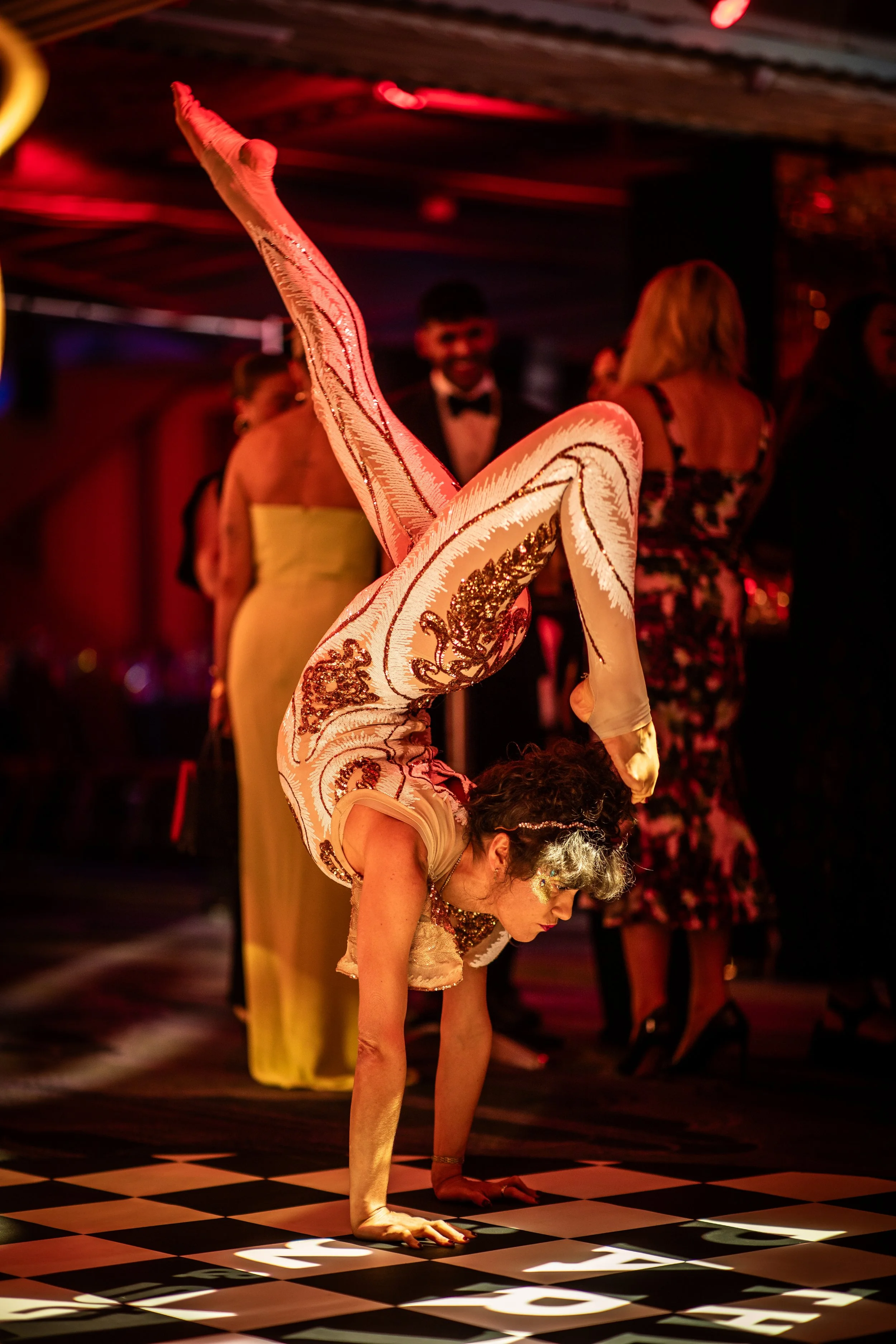 A woman performing a handstand on a checkered dance floor, with her legs extended upward and her head near the ground. There are other people in fancy attire in the background, in a dimly lit room with a red and warm lighting ambiance.