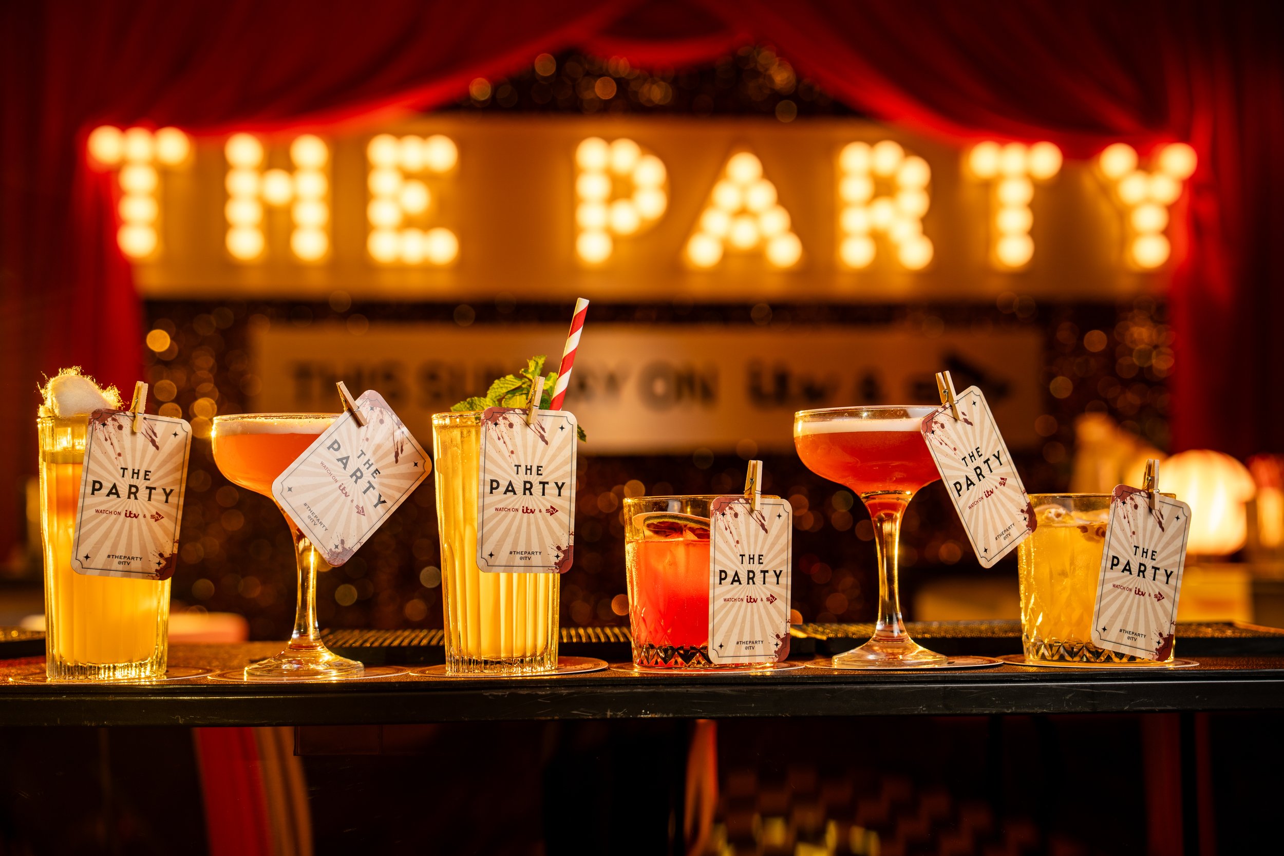 Several colorful cocktails with decorations and tags labeled "The Party" on a bar counter, with a lit marquee sign in the background that says "THEAT".