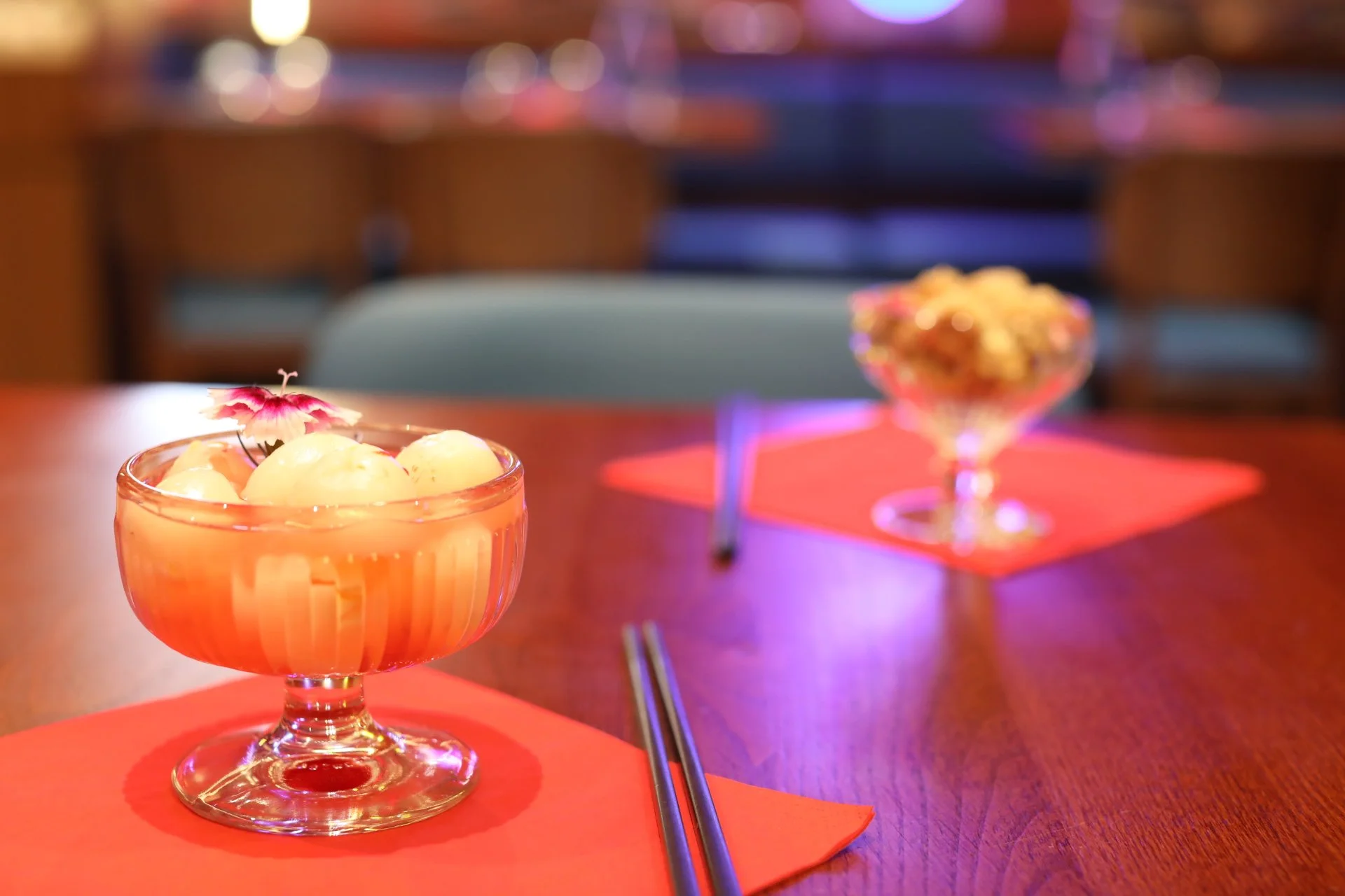 Un dessert dans un verre à pied avec une fleur décorative, sur une nappe rouge, en premier plan. En arrière-plan, un autre dessert dans un verre avec des fleurs colorées, dans un environnement éclairé et élégant.