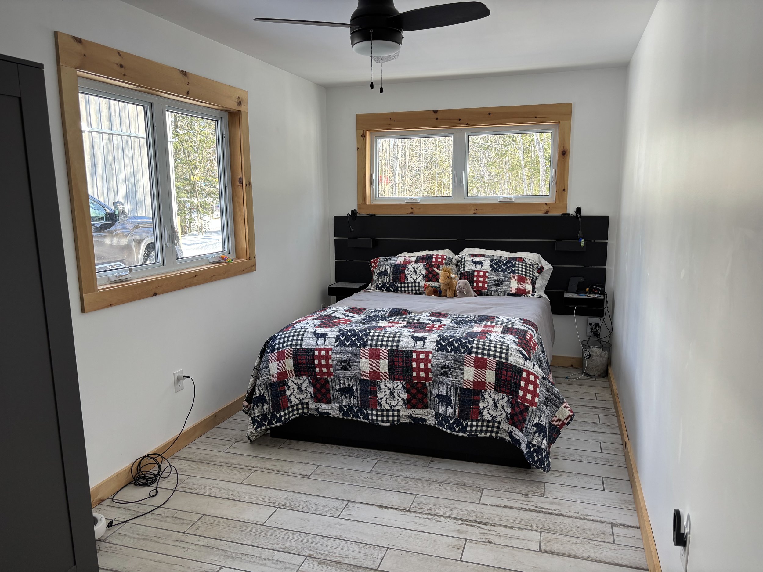 A cozy bedroom with a bed featuring a plaid quilt and matching pillows. The room has two large windows, a ceiling fan, and light wood trim. There are stuffed animals on the bed, and a power outlet with cords on the floor.