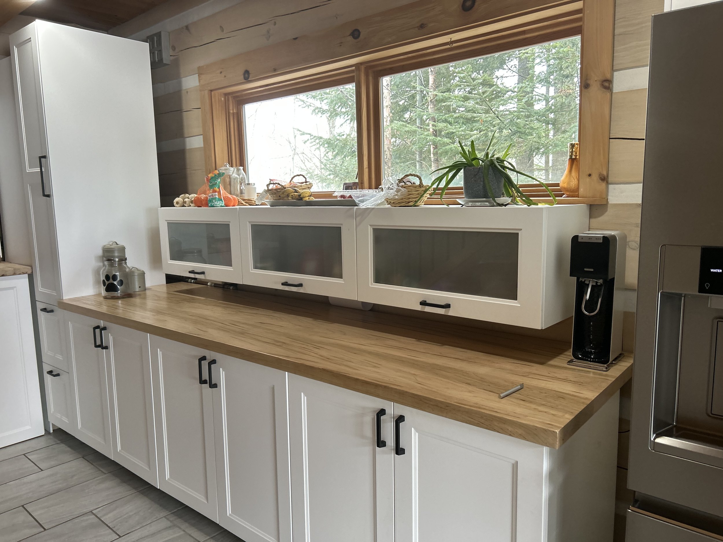 Modern kitchen with white pantry cabinets, wooden countertops, a large window, glass-paneled cabinet doors, and a stainless steel water dispenser.