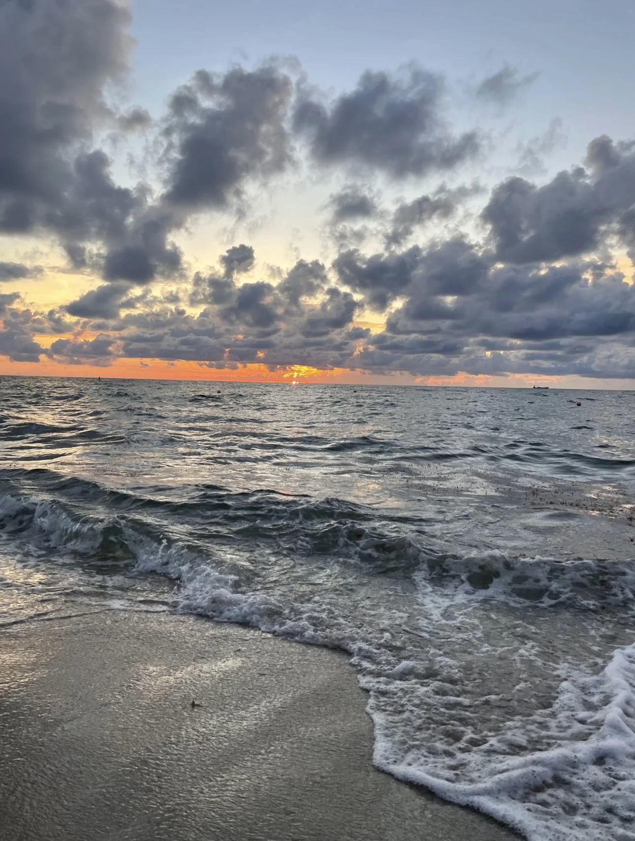 Sunset over the ocean with cloudy sky and gentle waves on the shore.