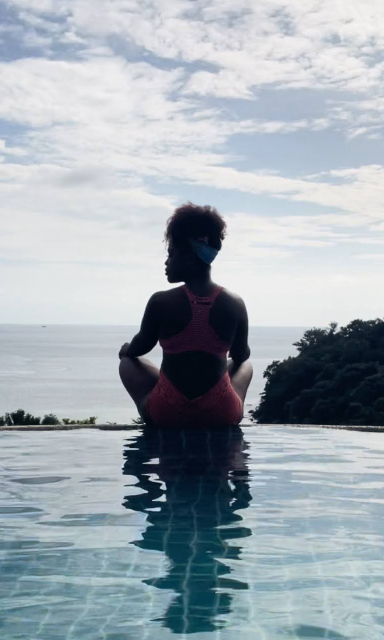 Silhouette of a woman sitting on the edge of a pool, overlooking the ocean, with a cloudy sky above and wearing a face mask and a swimsuit.