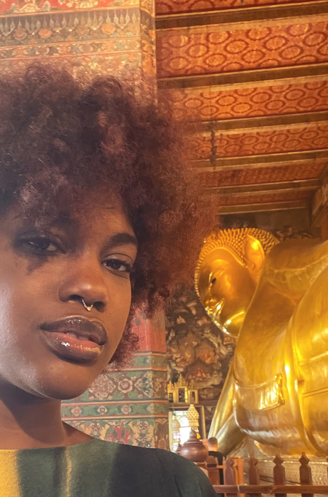 A woman with dark skin and curly hair stands in front of a large, reclining gold Buddha statue inside a temple with ornate, colorful wall decorations and gold accents.