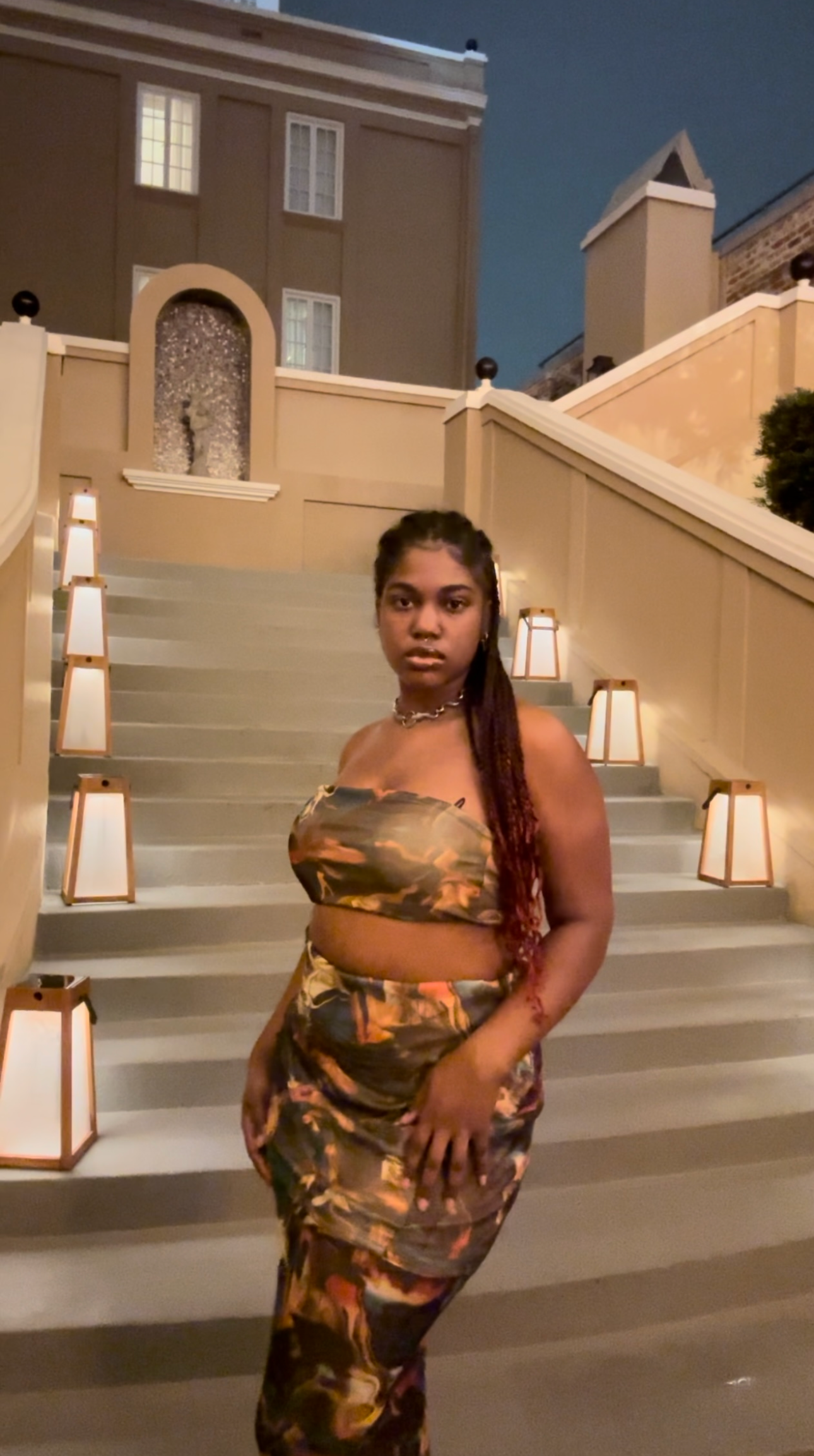 A woman in a matching camouflage outfit standing on stairs at night, with decorative lamps lining the sides and a building in the background.