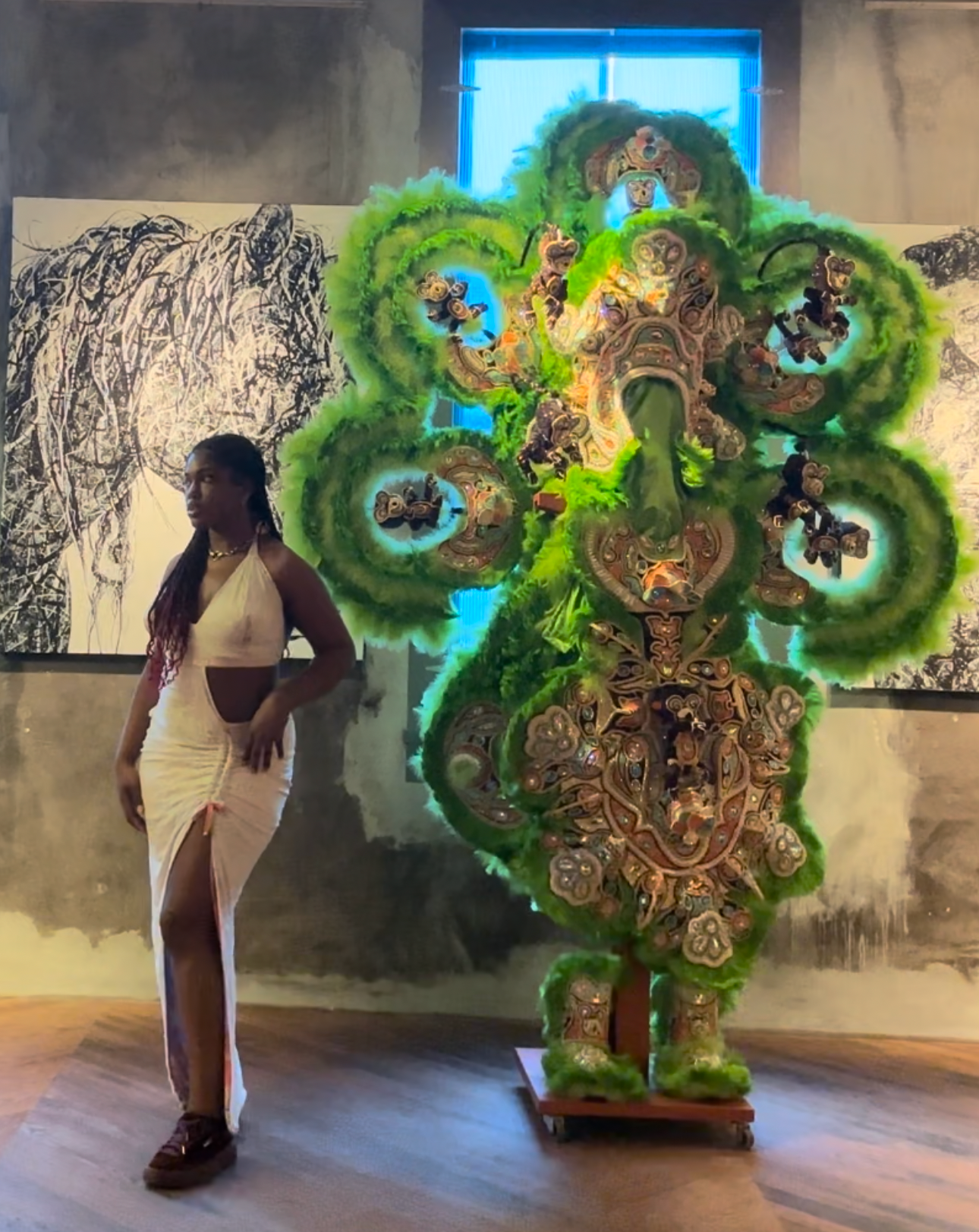 A woman in a white dress standing next to an elaborate, green and multicolored tapestry or sculpture with intricate designs, displayed indoors with artwork visible on the walls.