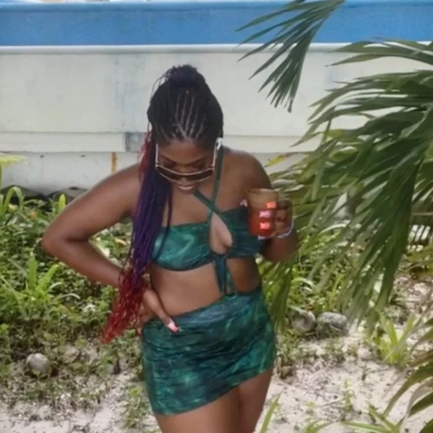 Woman in green tie-dye dress with purple and red braids, wearing sunglasses, holding a drink, outdoors near plants and sand.