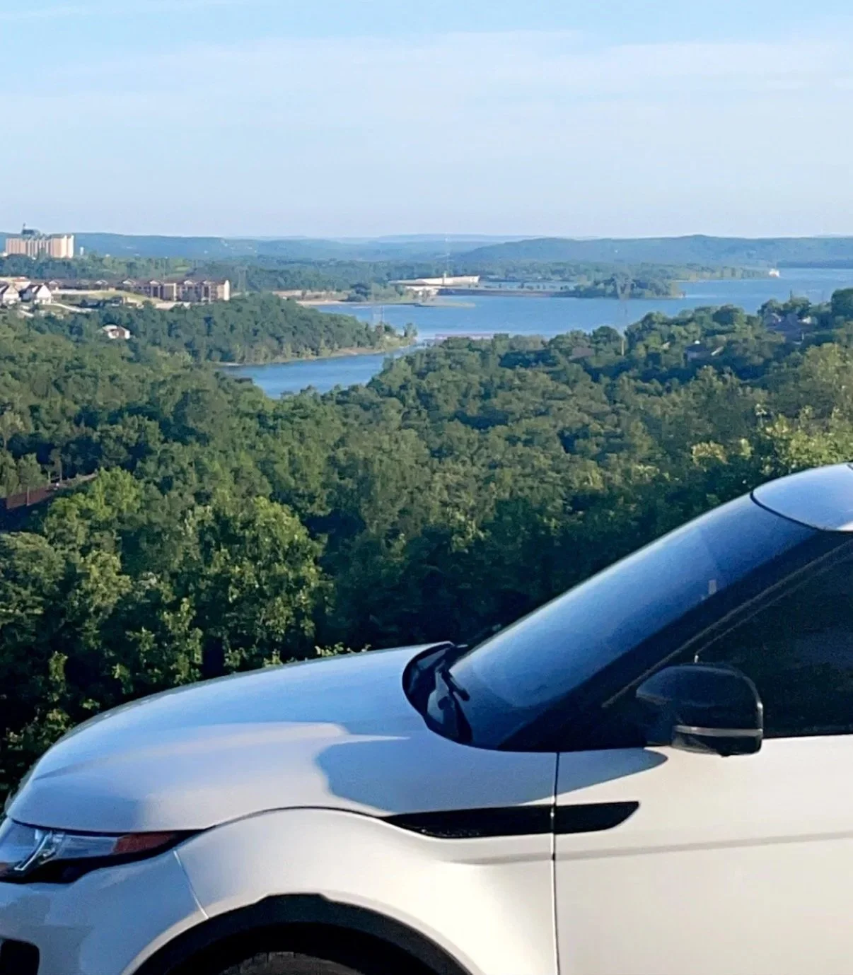 Partial view of a white car parked in front of a scenic landscape with a river, green trees, and distant buildings under a blue sky.