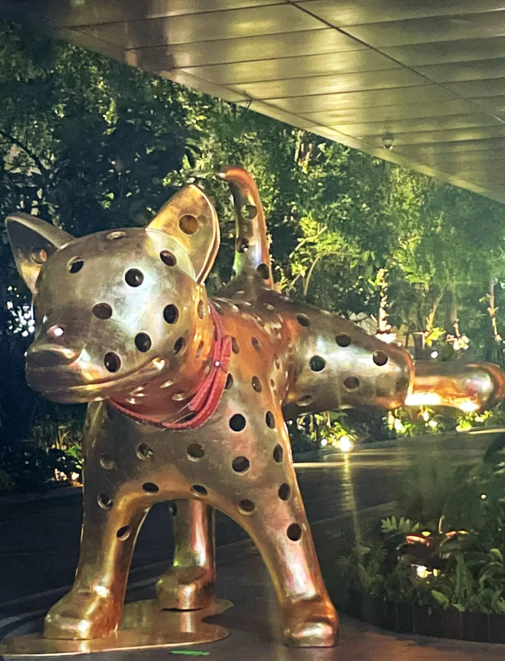 A metallic sculpture of a dog with circular holes all over its body, wearing a red collar, situated outdoors with greenery and a building’s overhang in the background.