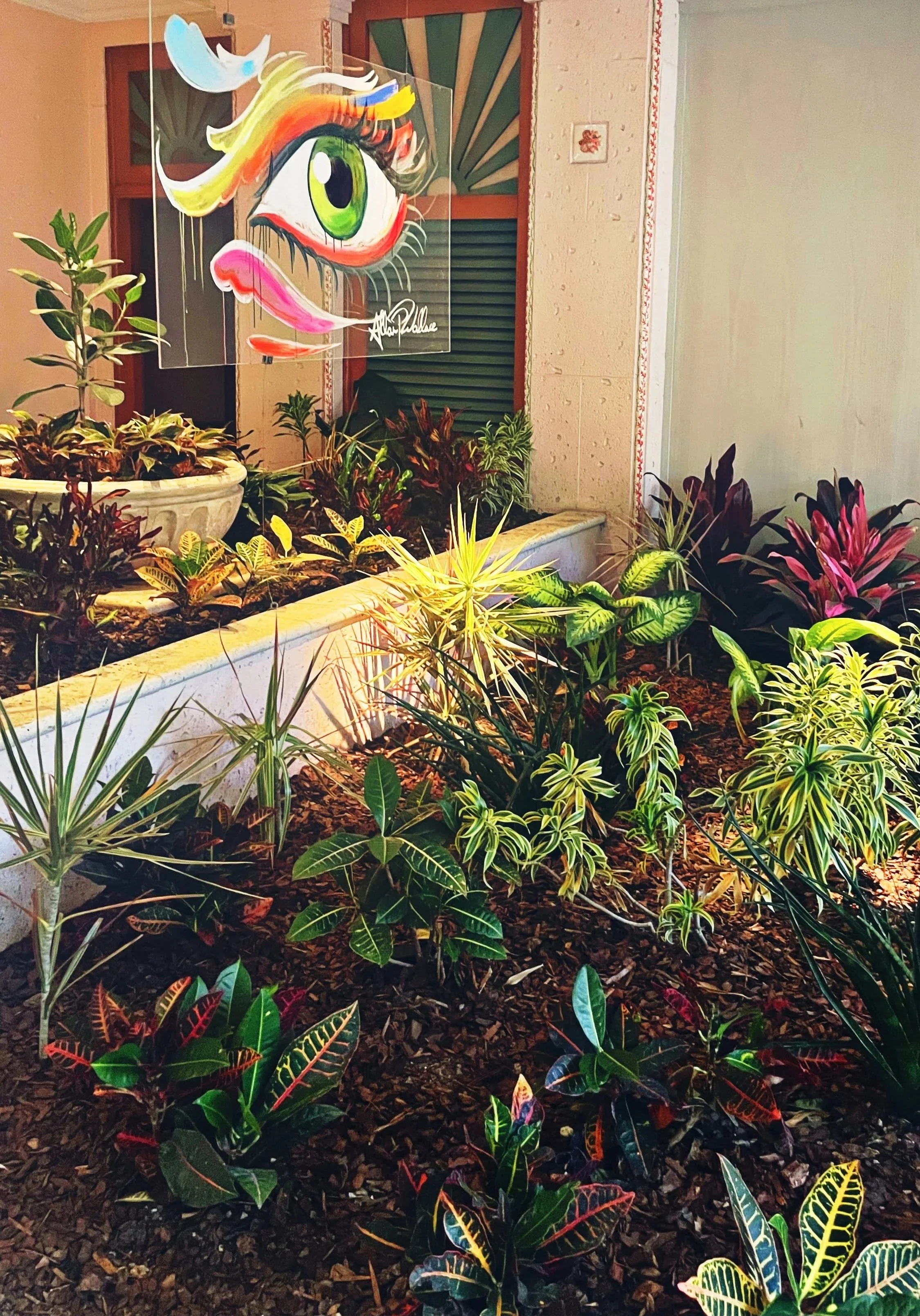 A garden bed with a variety of colorful plants and leaves, in front of a wall with a window. Hanging on the wall is a large, colorful painting of a cartoonish eye with vibrant makeup, making a playful expression.