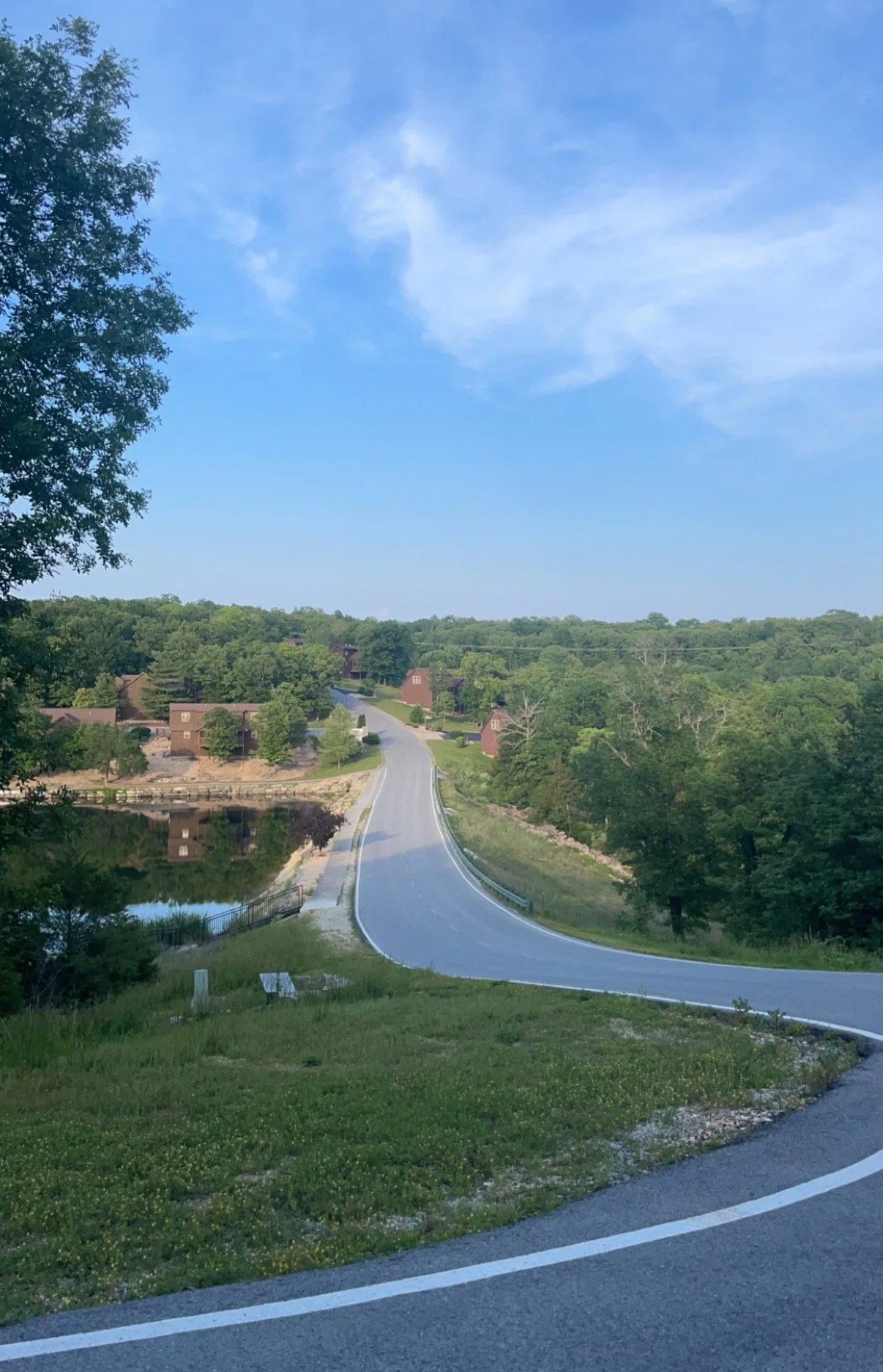 Curving road in a rural area with green trees, houses, and a pond reflecting the sky and trees, under a partly cloudy blue sky.
