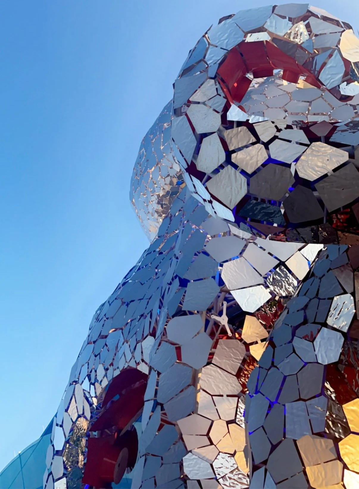 Close-up of a modern architectural structure made of reflective, irregularly shaped mirrors with a clear blue sky in the background.