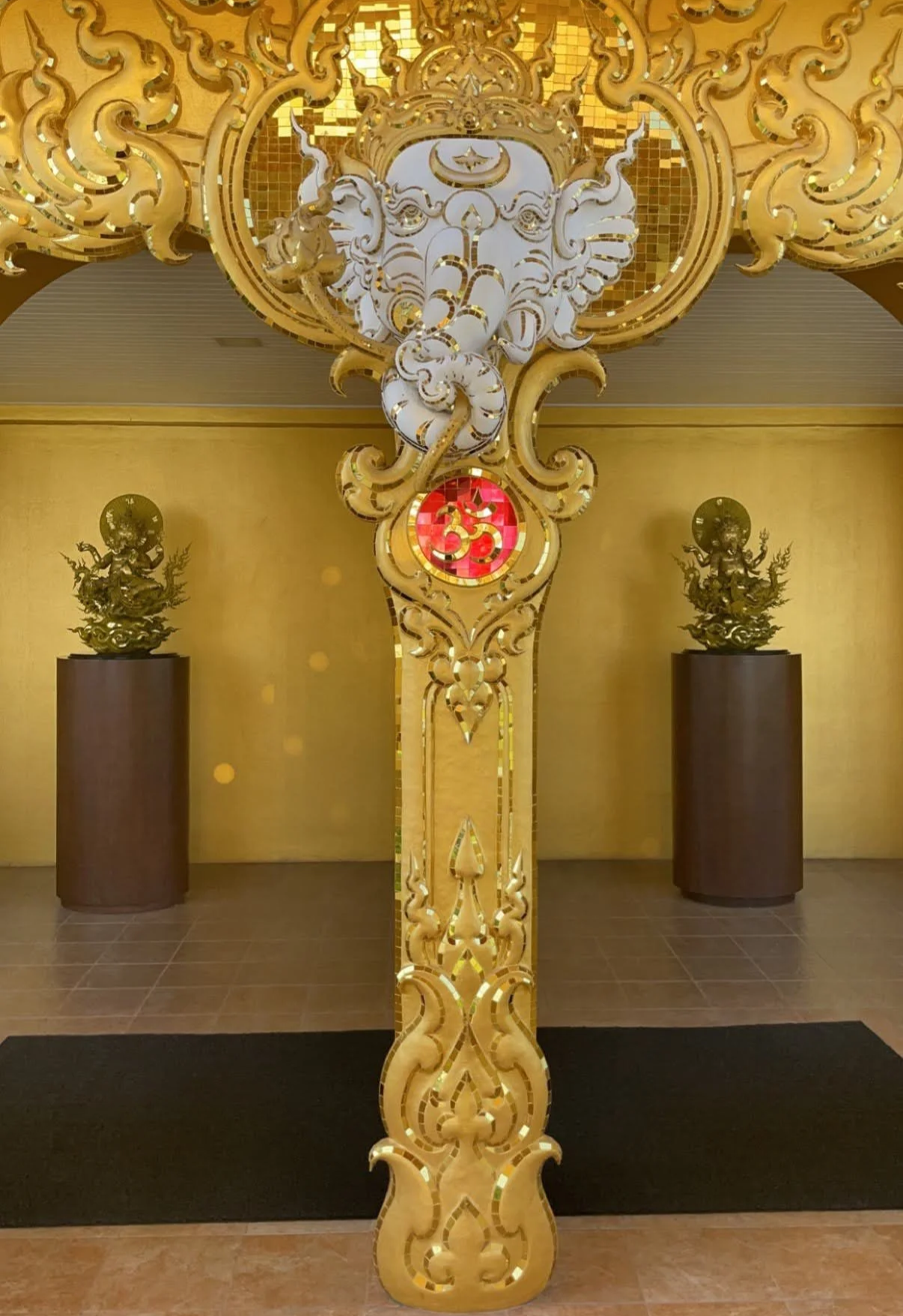 Ornate gold and white decorative structure featuring an elephant head with multiple trunks and an 'Om' symbol in red, set against a gold background with two smaller gold statues on pedestals.