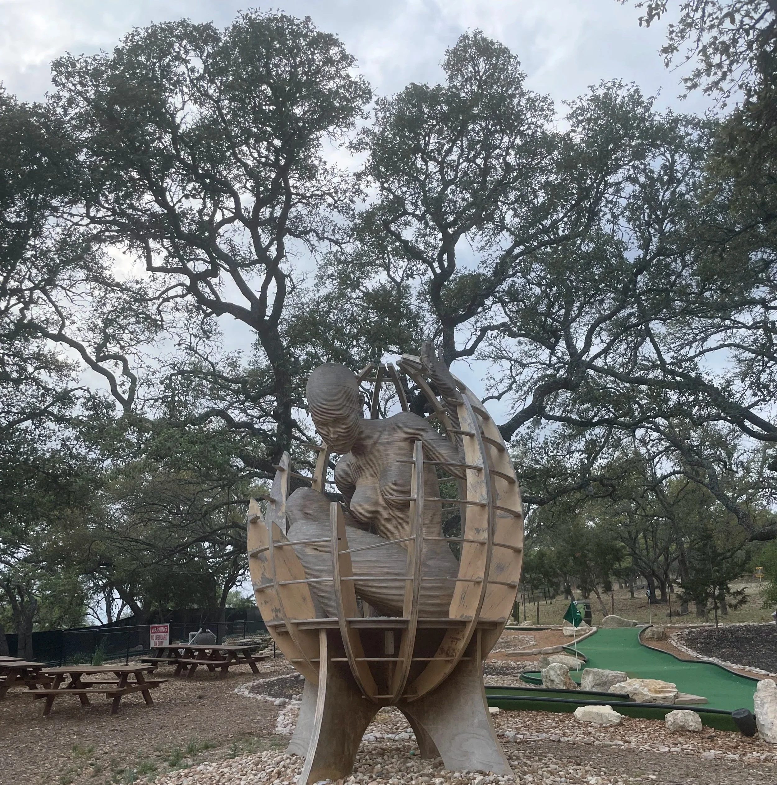 A wooden sculpture of a fetus inside a globe, set in a park with trees and a mini golf course in the background.