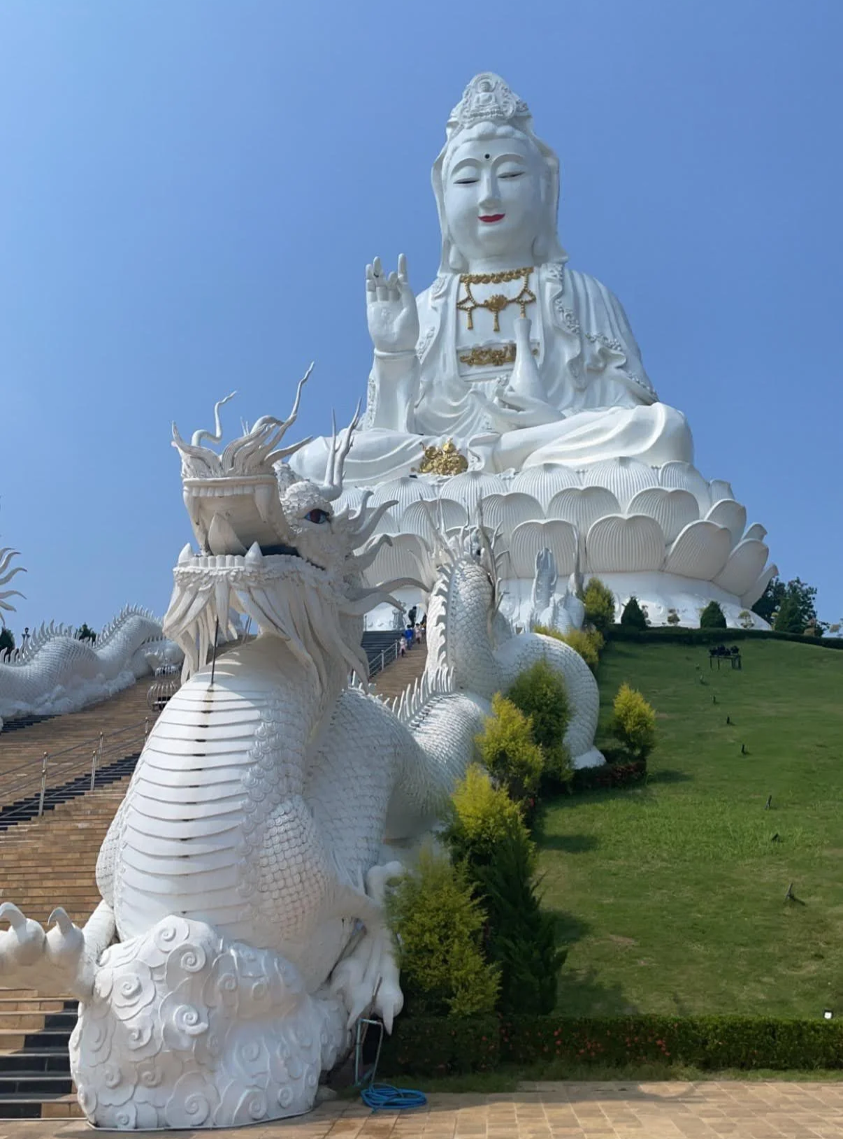 A large white statue of a female deity seated on a lotus flower, with a dragon sculpture in front, against a clear blue sky.