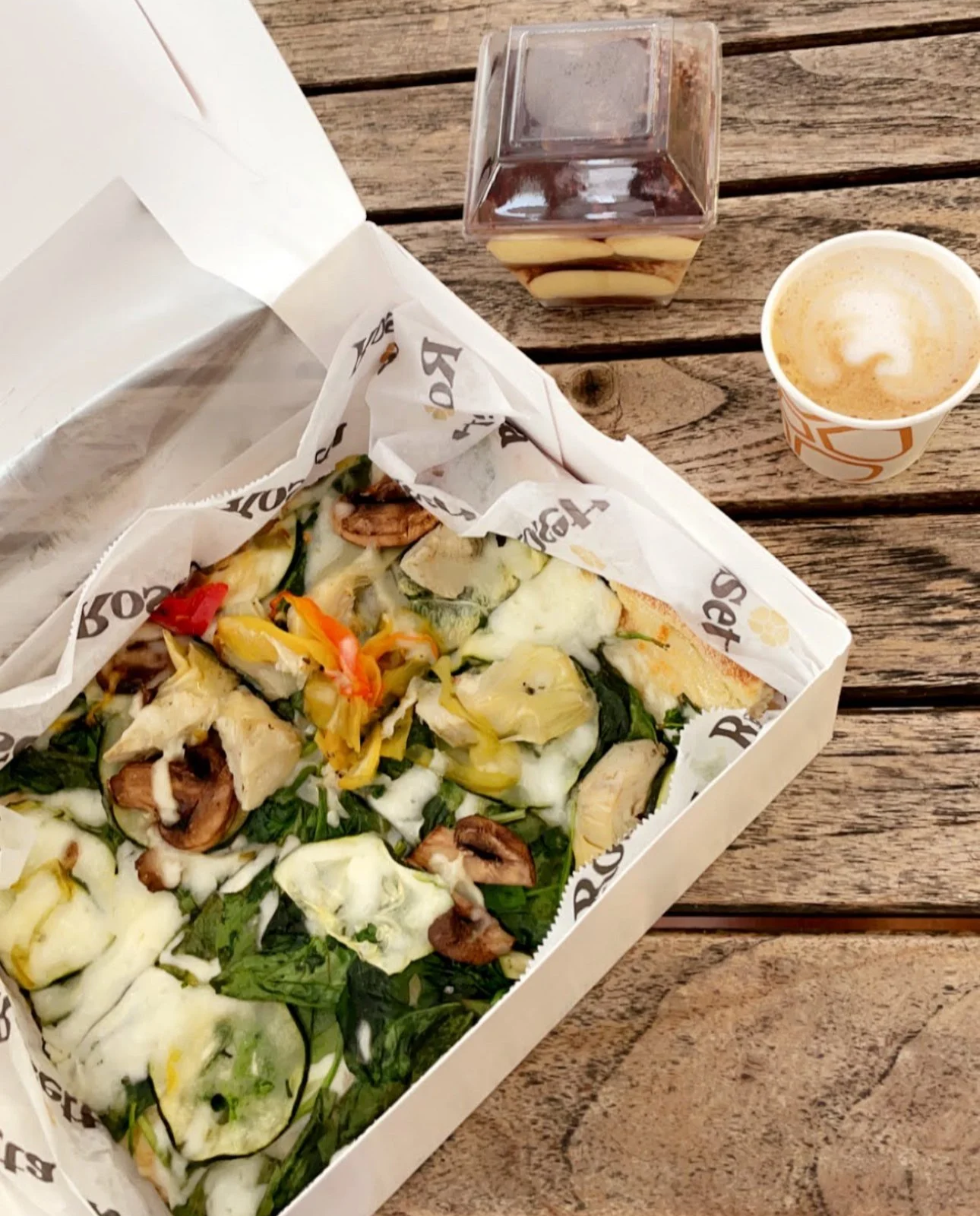 A pizza with spinach, mushrooms, and peppers in a takeout box on a wooden table, with a glass of iced tea and a cup of frothy coffee nearby.