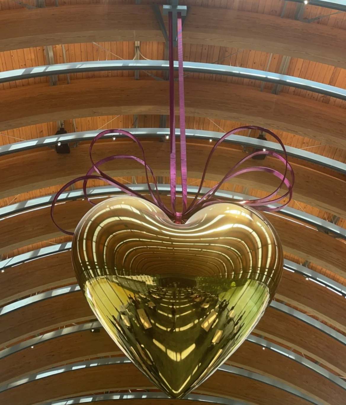 A large, shiny gold heart-shaped sculpture hanging from a purple ribbon inside a building with a wooden ceiling and curved architectural features.