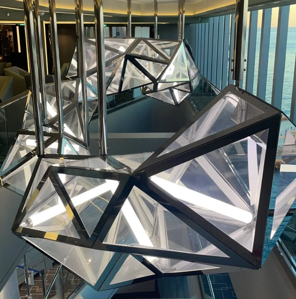Modern geometric chandelier with illuminated triangular glass panels hanging indoors with large windows showing ocean views.