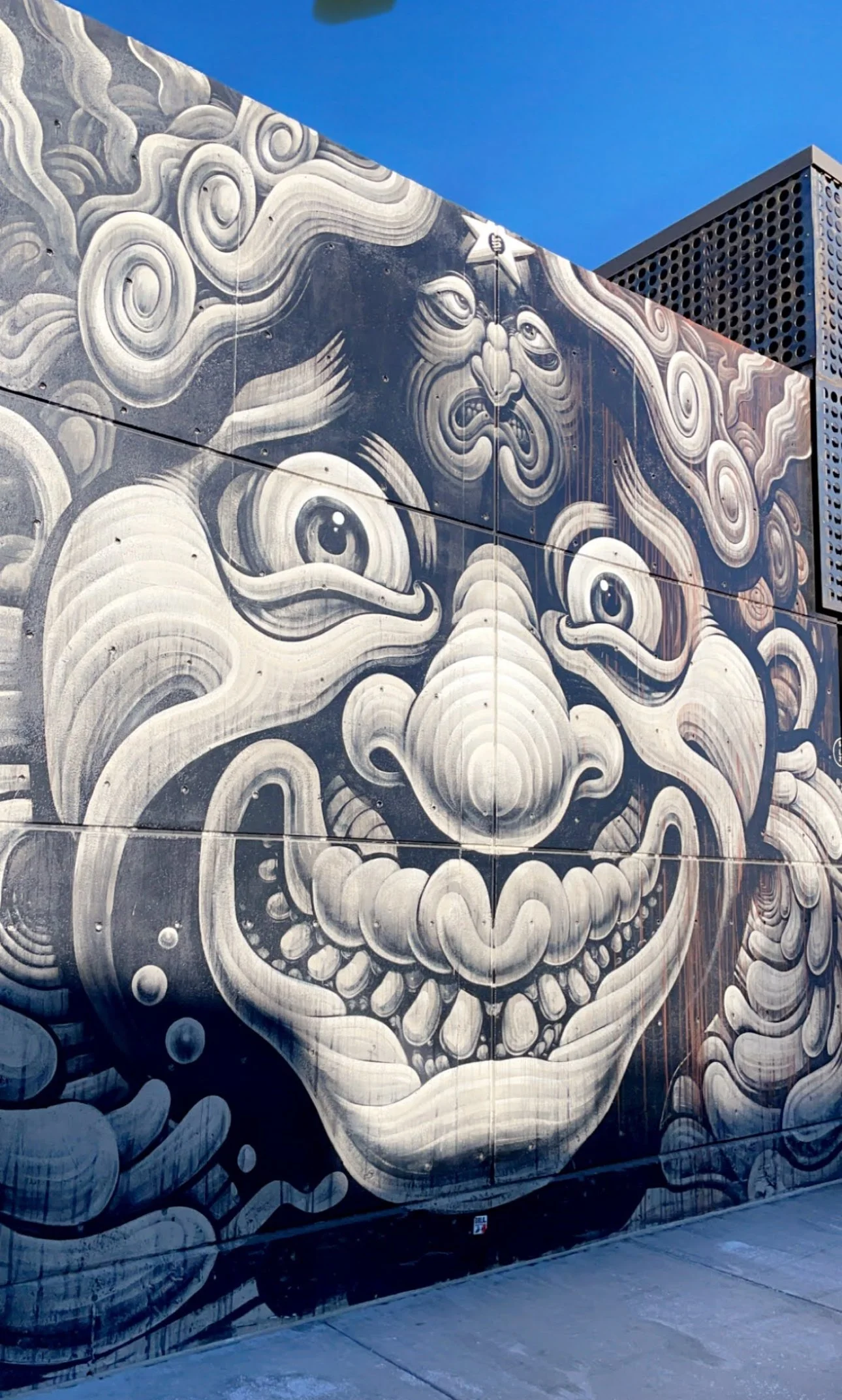 Street art mural featuring a surreal, monochrome face with large eyes, exaggerated nose, wide grin showing many teeth, and swirling patterns surrounding the face on an exterior wall against a blue sky.