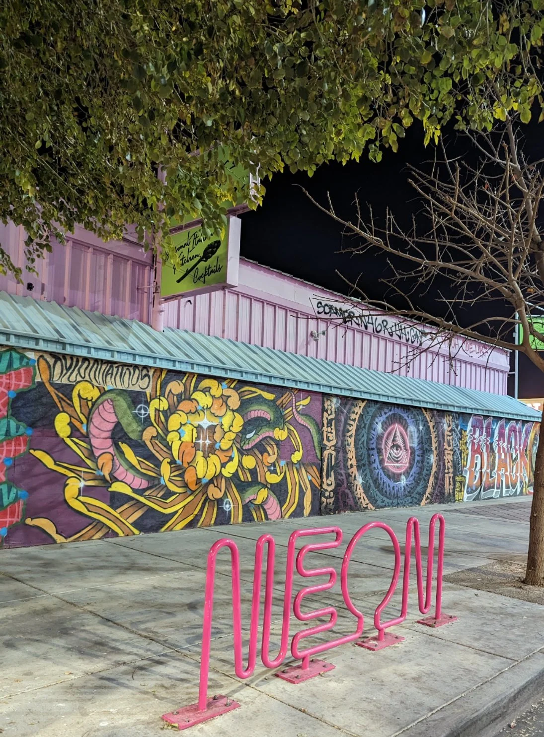 Night scene featuring a pink mural wall with colorful graffiti art, a green sign for a bar, leafless trees, and a bright pink neon sign spelling MUSEUM on the sidewalk.