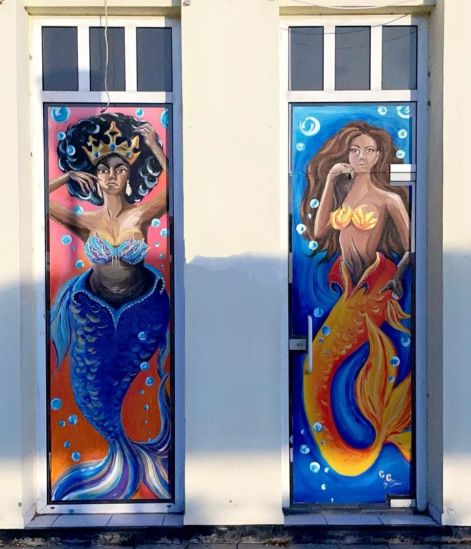 Colorful mural painting on two doors, depicting two women with fish tails, mermaids, surrounded by bubbles. The left mermaid has dark curly hair, a crown, and a blue tail; the right mermaid has long brown hair and a yellow-orange tail.